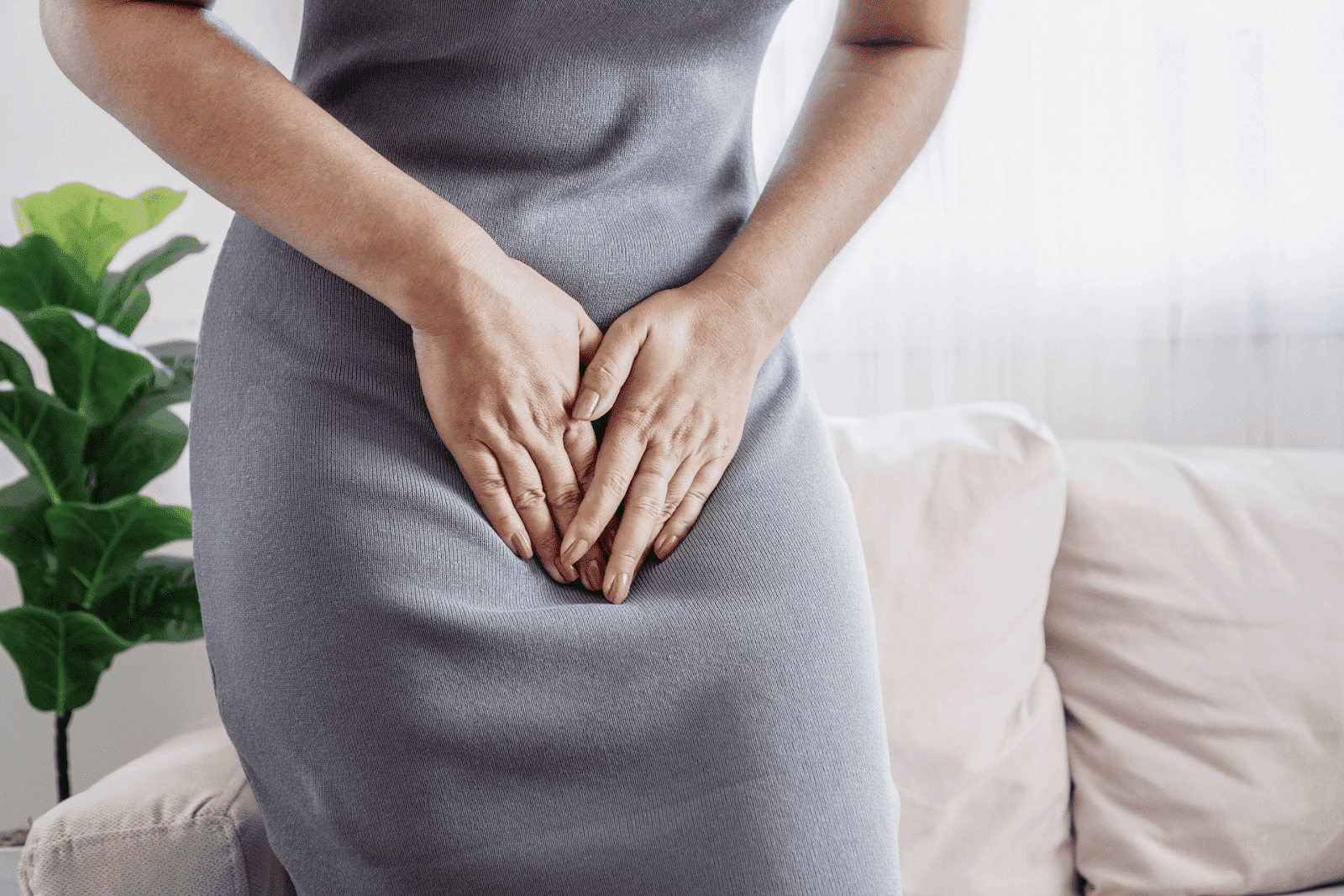 A person standing indoors with hands clasped over their pelvic area, suggesting vaginal or urinary discomfort.