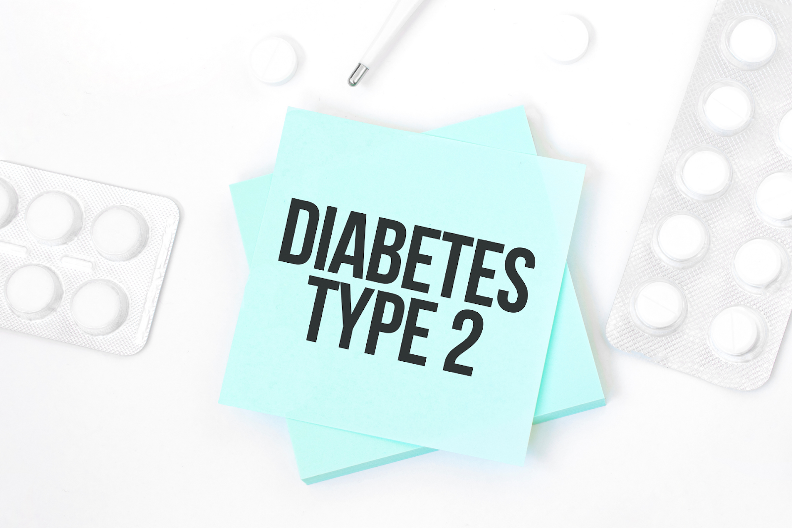 A blue sticky note reading ‘DIABETES TYPE 2’ surrounded by blister packs of white tablets and a thermometer.