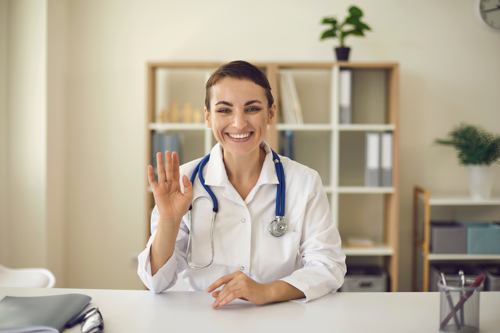 A cheerful doctor with a stethoscope waves while sitting at a desk in a bright office.