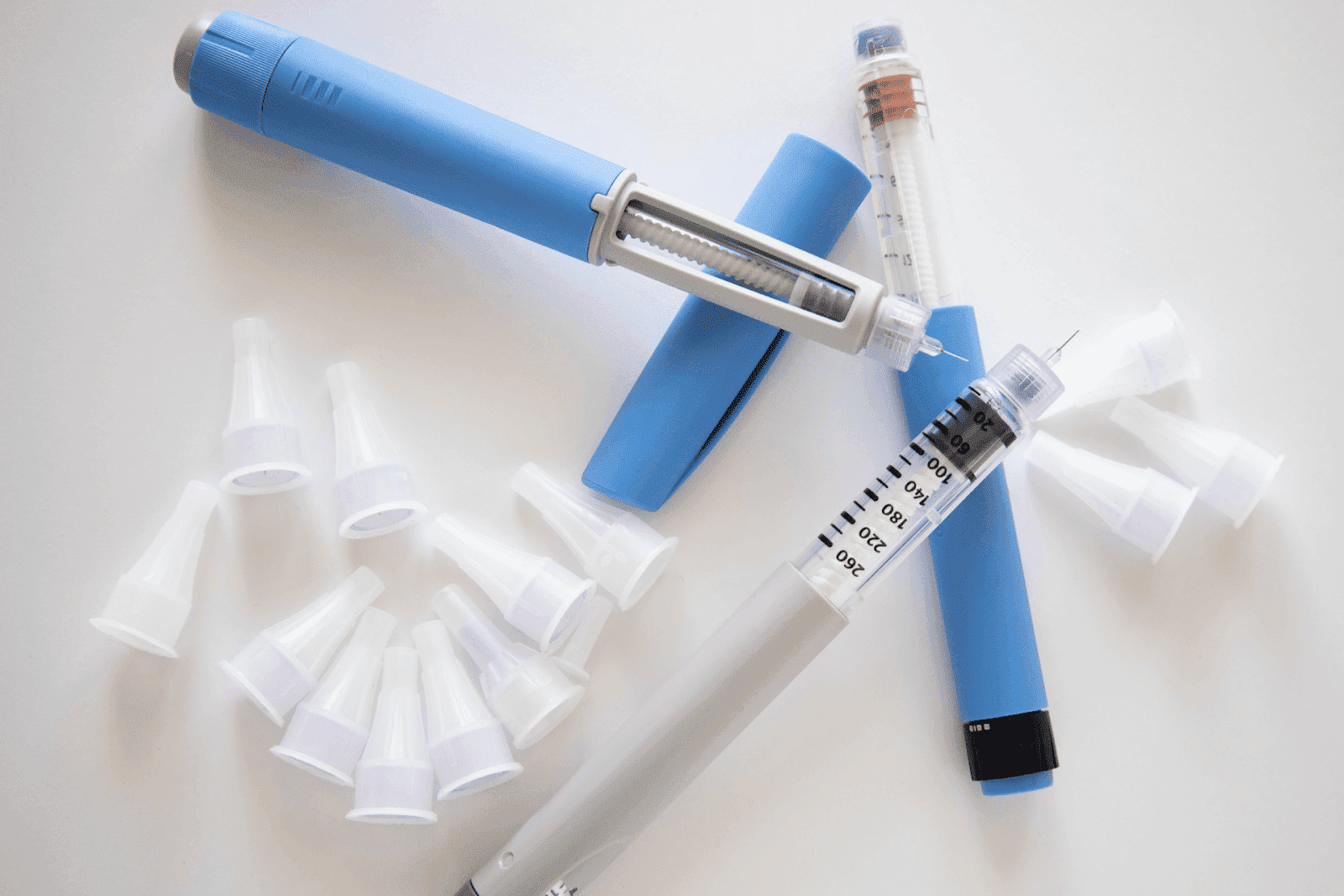 A close-up image showing two insulin pens, one in blue and the other in grey, with clear dosage windows and needles attached, surrounded by multiple white disposable pen needle caps on a white surface.