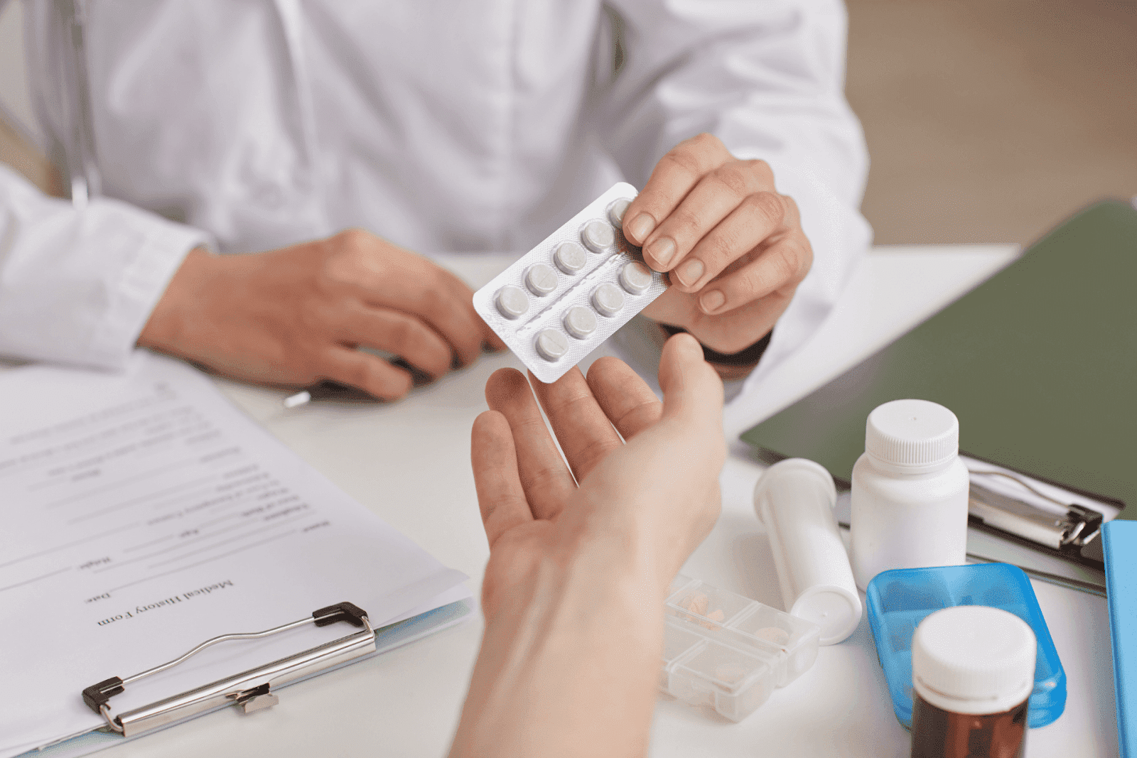 A doctor handing a patient a blister pack of pills across a desk with medical forms and medication bottles