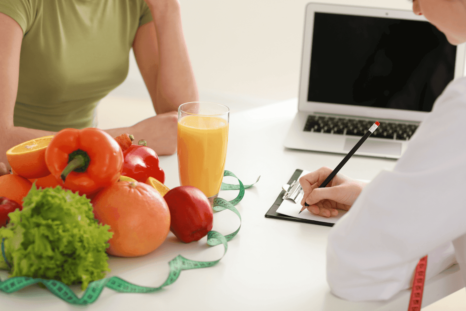A doctor takes notes while sitting across from a patient with fresh fruits, vegetables, and juice on the table.