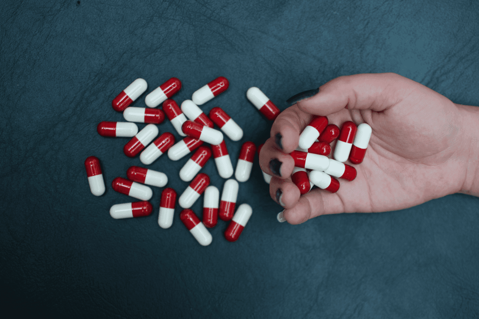 A hand holding several red-and-white capsules with more capsules scattered on a dark surface.