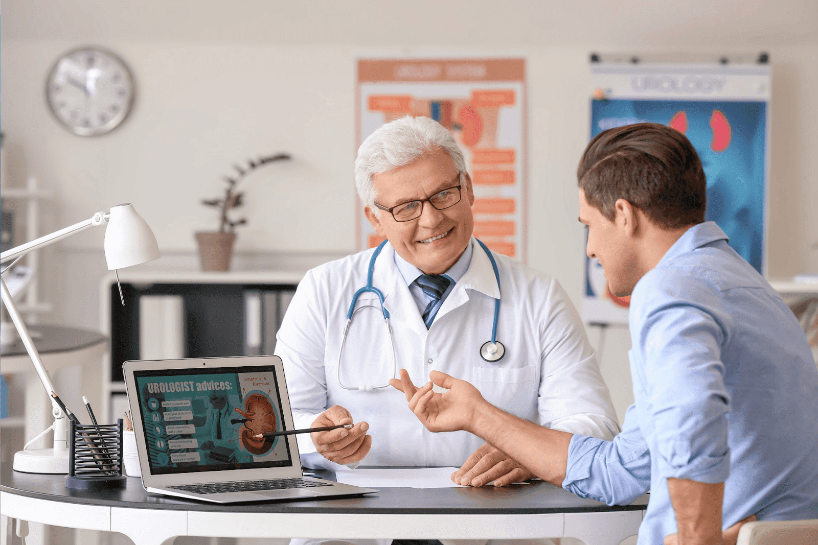 A male patient consults with a smiling urologist who is pointing at a kidney diagram on a laptop screen.