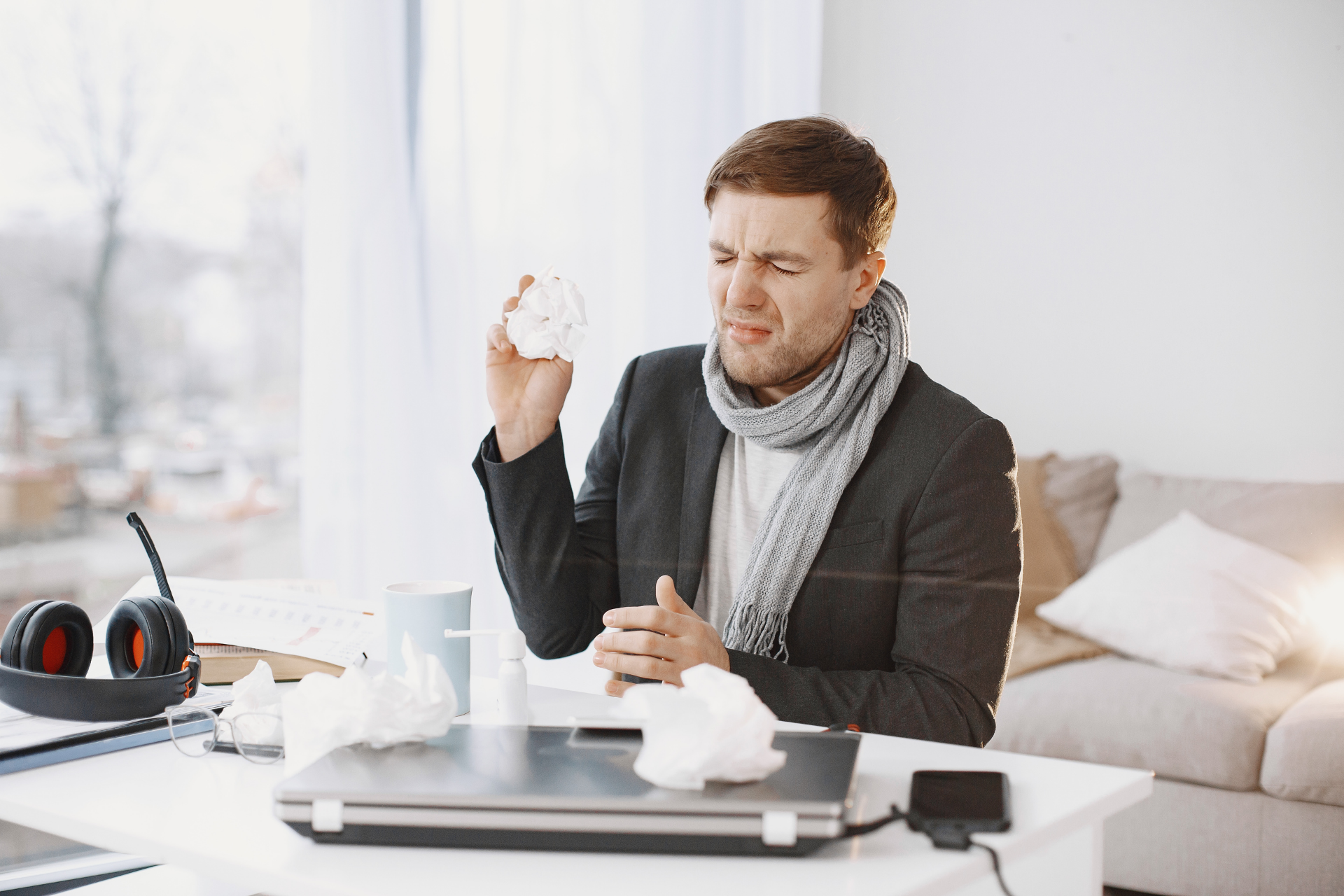 A man at a desk sneezes into a tissue, with a laptop and scattered tissues in front of him.