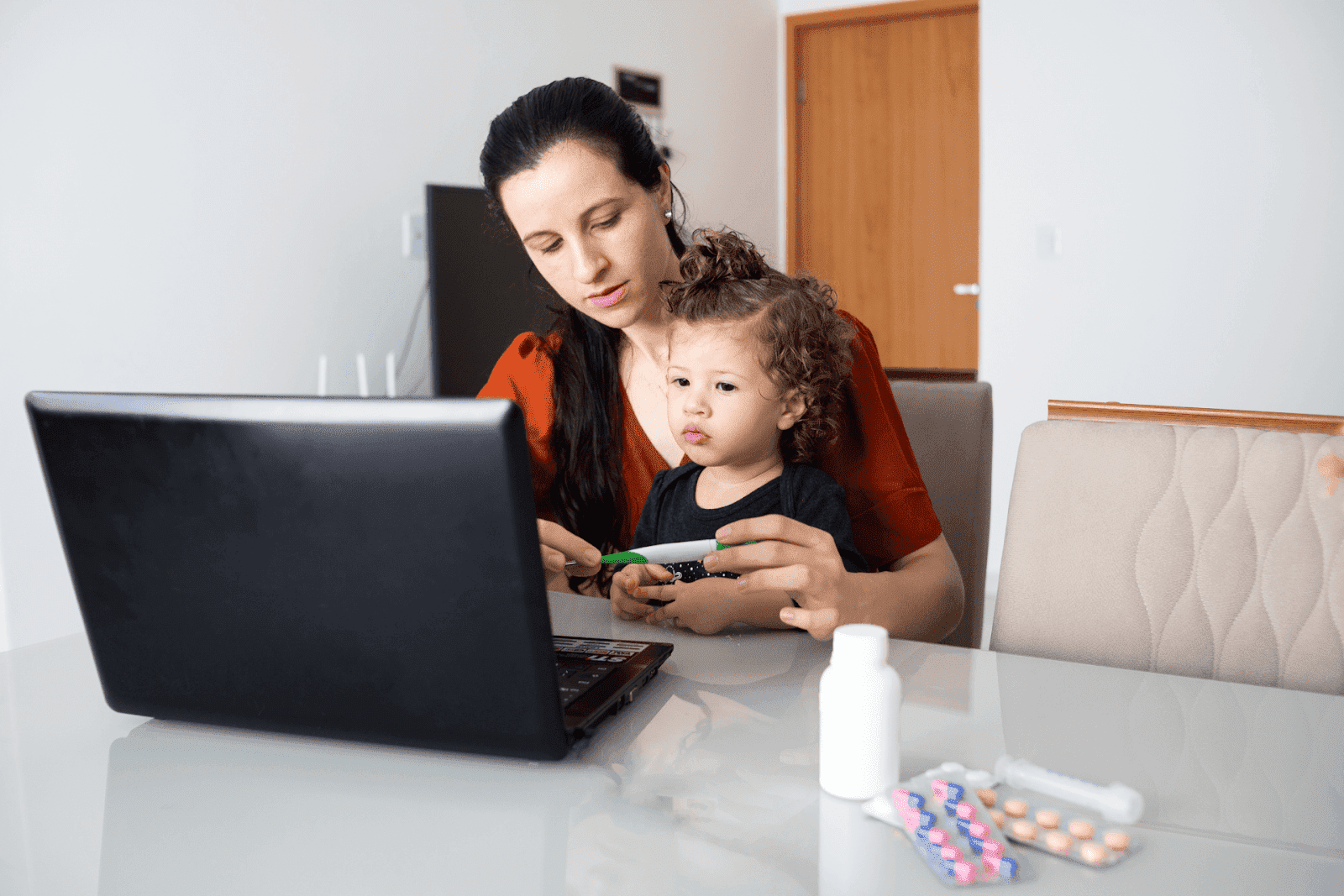 A mother and child sit at a table, showing a thermometer during an online doctor visit