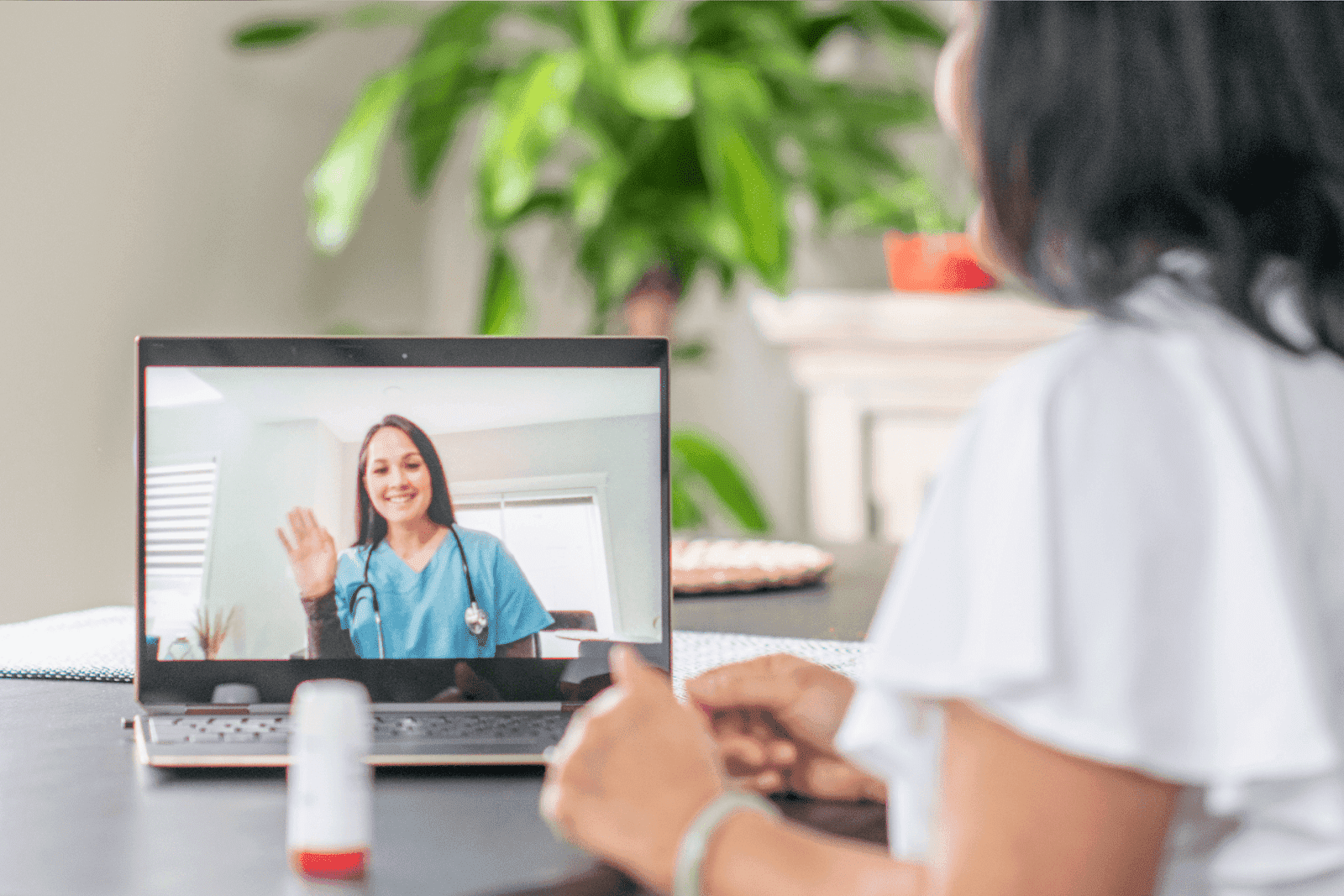 A patient on a laptop video call with a smiling doctor in blue scrubs waving.