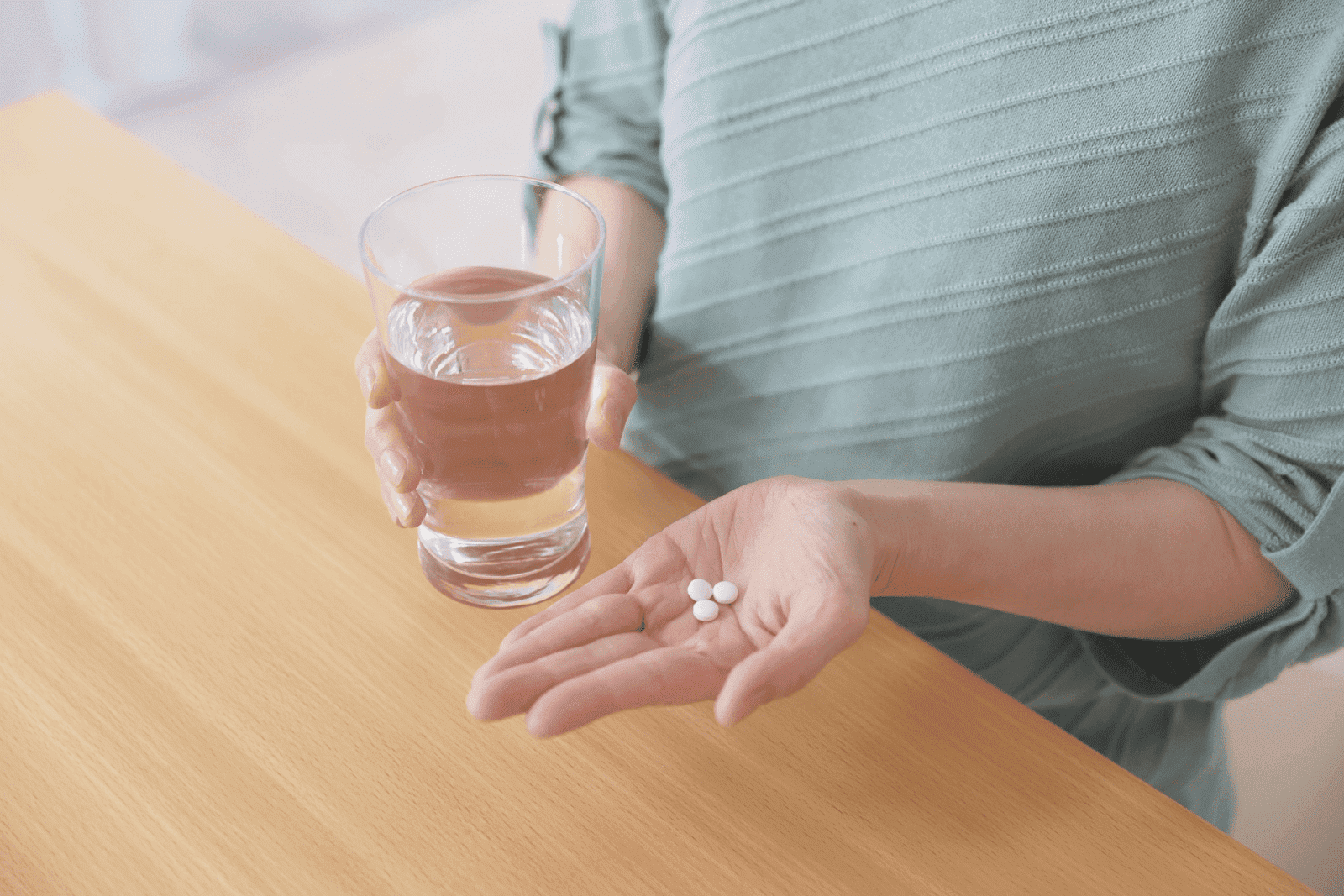 A person holding three white tablets in one hand and a glass of water in the other.