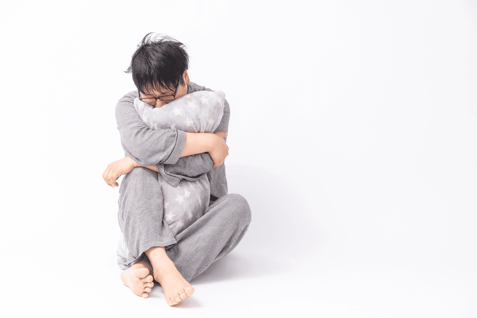 A person sitting on the floor against a white background, hugging a pillow tightly with their head lowered, conveying sadness, anxiety, or emotional distress.