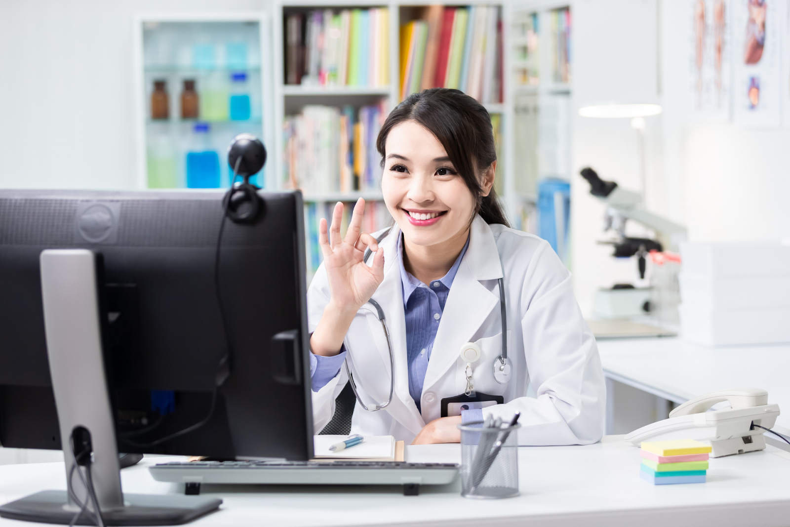 A smiling doctor in a white coat waves at a computer screen during a virtual consultation.