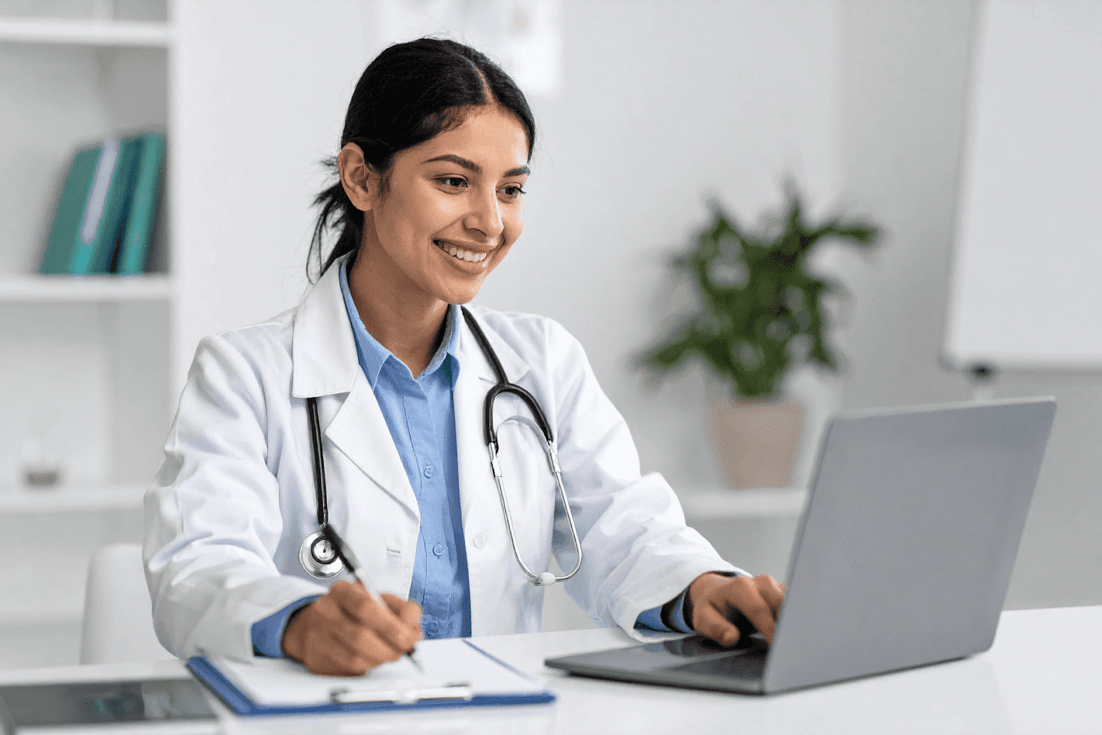 A smiling doctor sits at a desk with a laptop and clipboard, appearing to conduct an online consultation or enter patient notes.