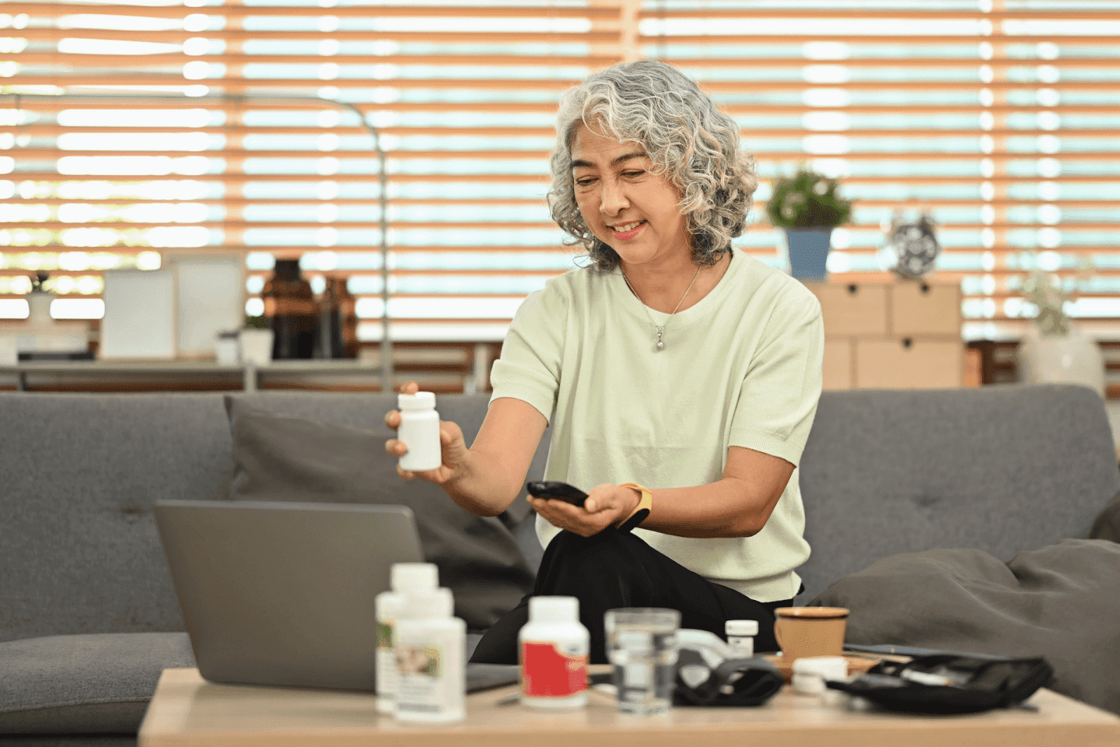 A smiling older woman holds up a pill bottle while sitting on a couch, showing it during a video call