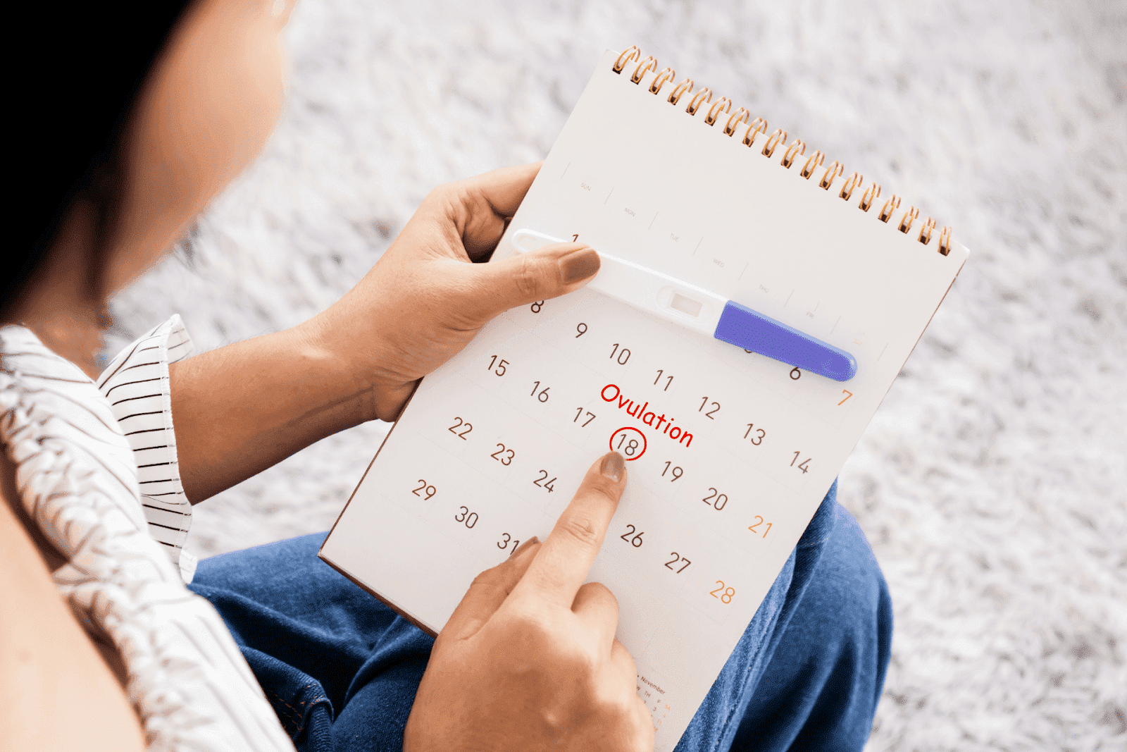 A woman points to the 18th marked as "Ovulation" on a calendar while holding a pregnancy test.