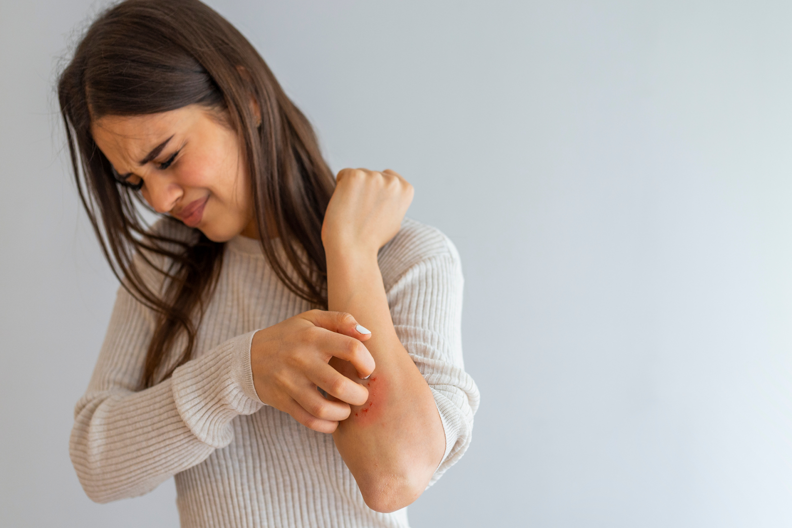 A woman scratching a red, irritated rash on her forearm and wincing in discomfort.
