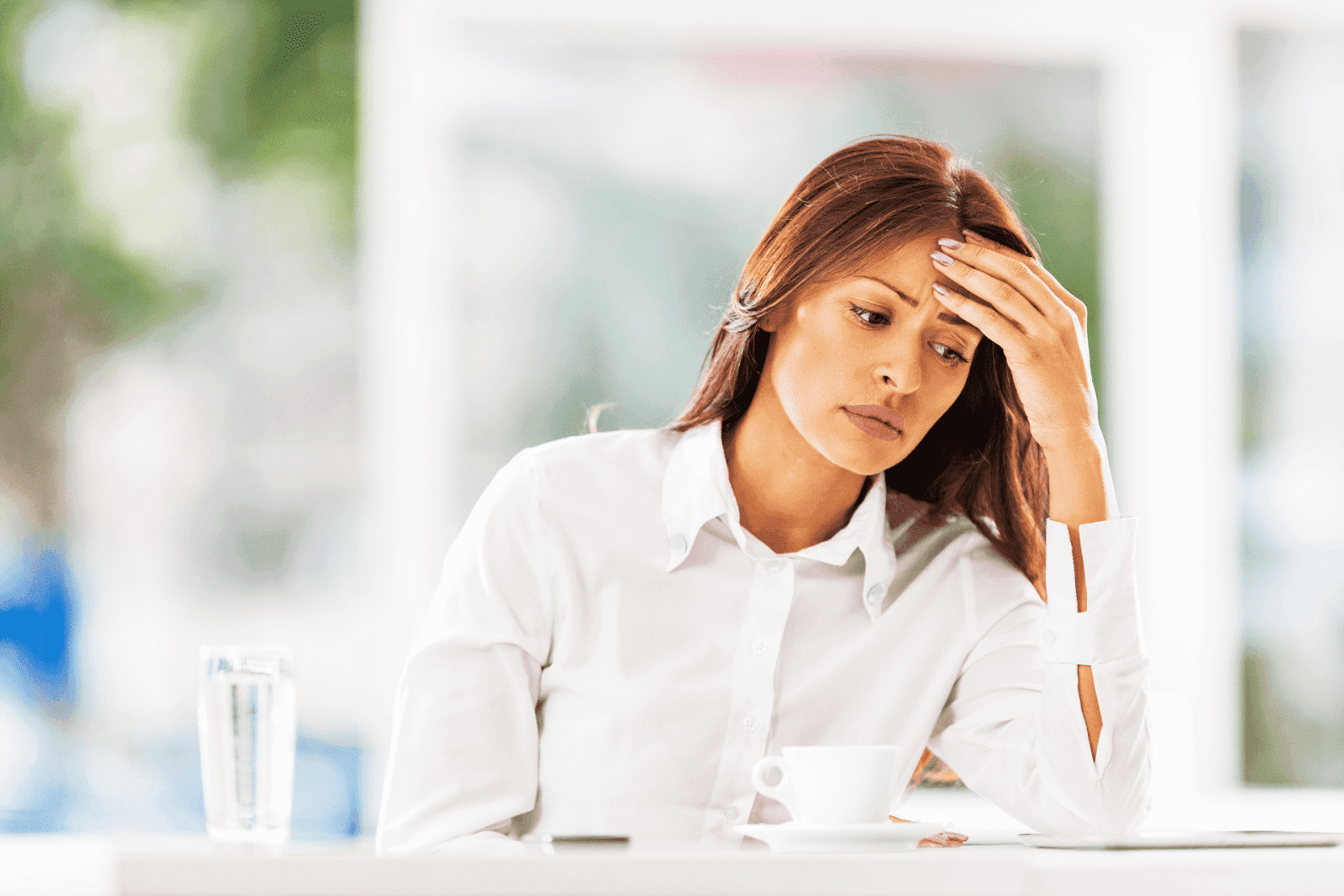 A woman sits at a table, looking stressed or fatigued, resting her head in her hand