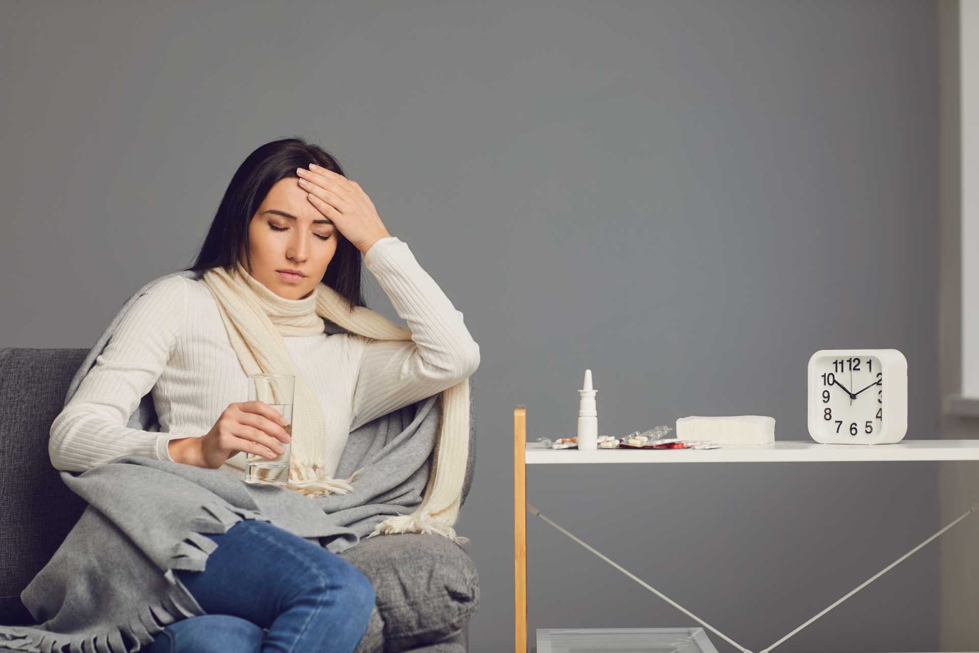 A woman sits on a chair holding a glass of water and touching her forehead, with medicine and a clock on a nearby table