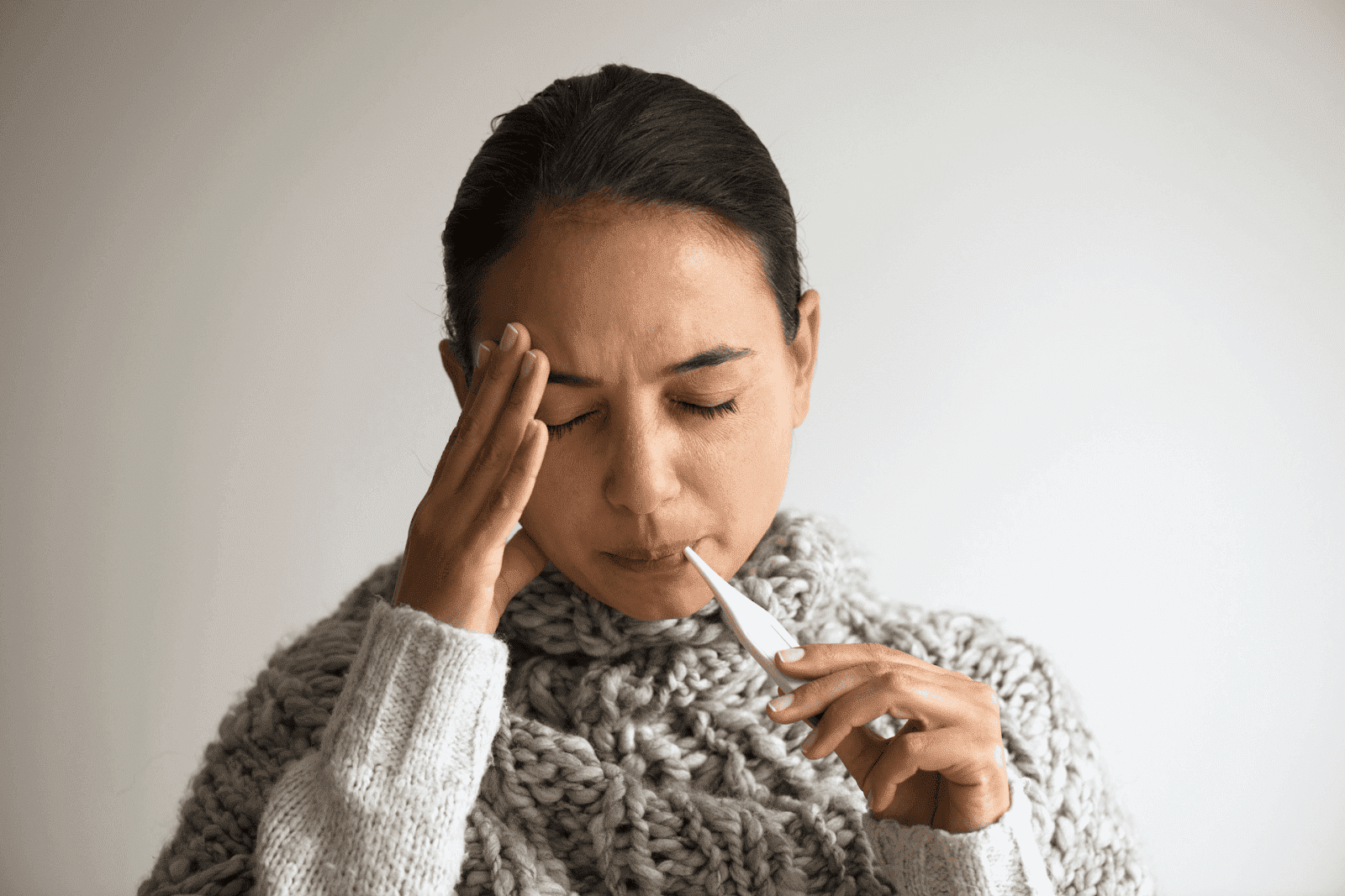 A woman wearing a thick knitted sweater holds a thermometer in her mouth while pressing her hand to her temple, eyes closed, appearing unwell against a neutral background.