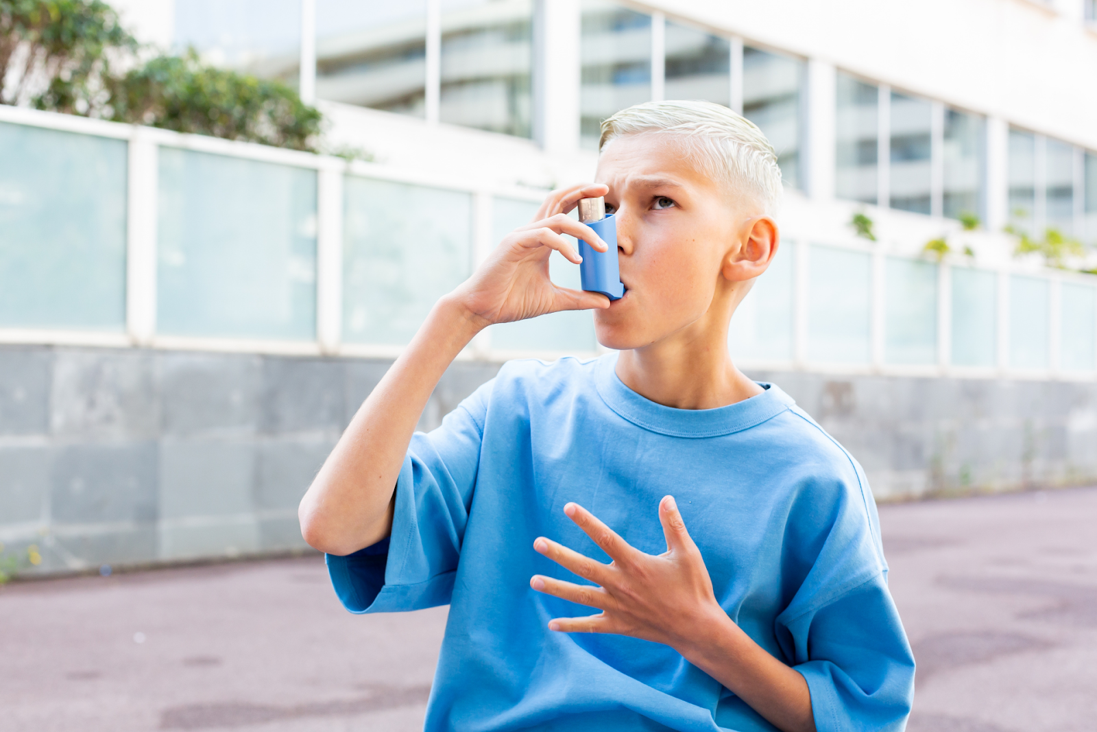 A young boy holding a blue inhaler to his mouth and clutching his chest, appearing to have trouble breathing.