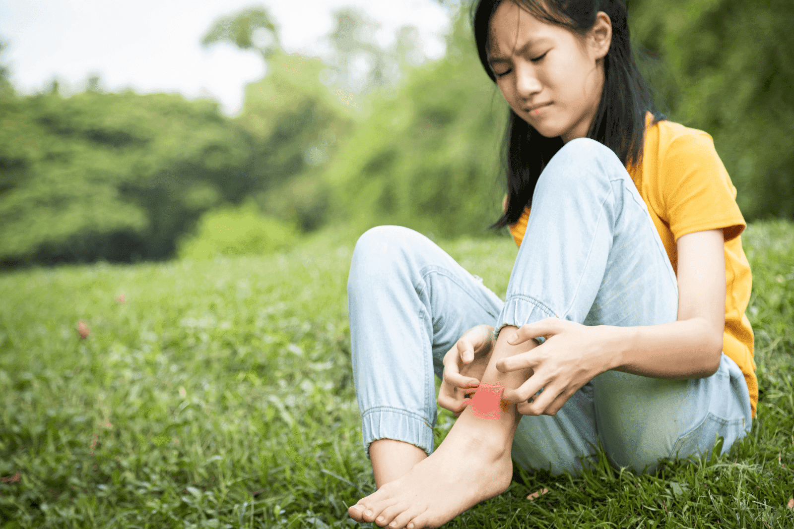A young girl sitting on grass outdoors, scratching her ankle with redness visible on her skin.