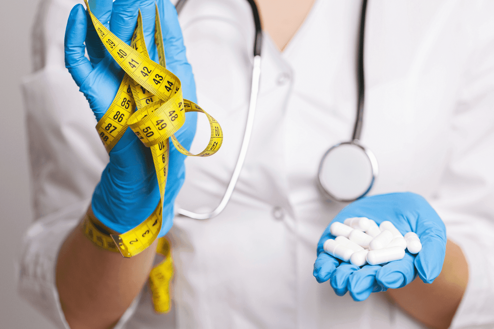 Close-up of a doctor wearing gloves holding white capsules in one hand and a yellow measuring tape in the other