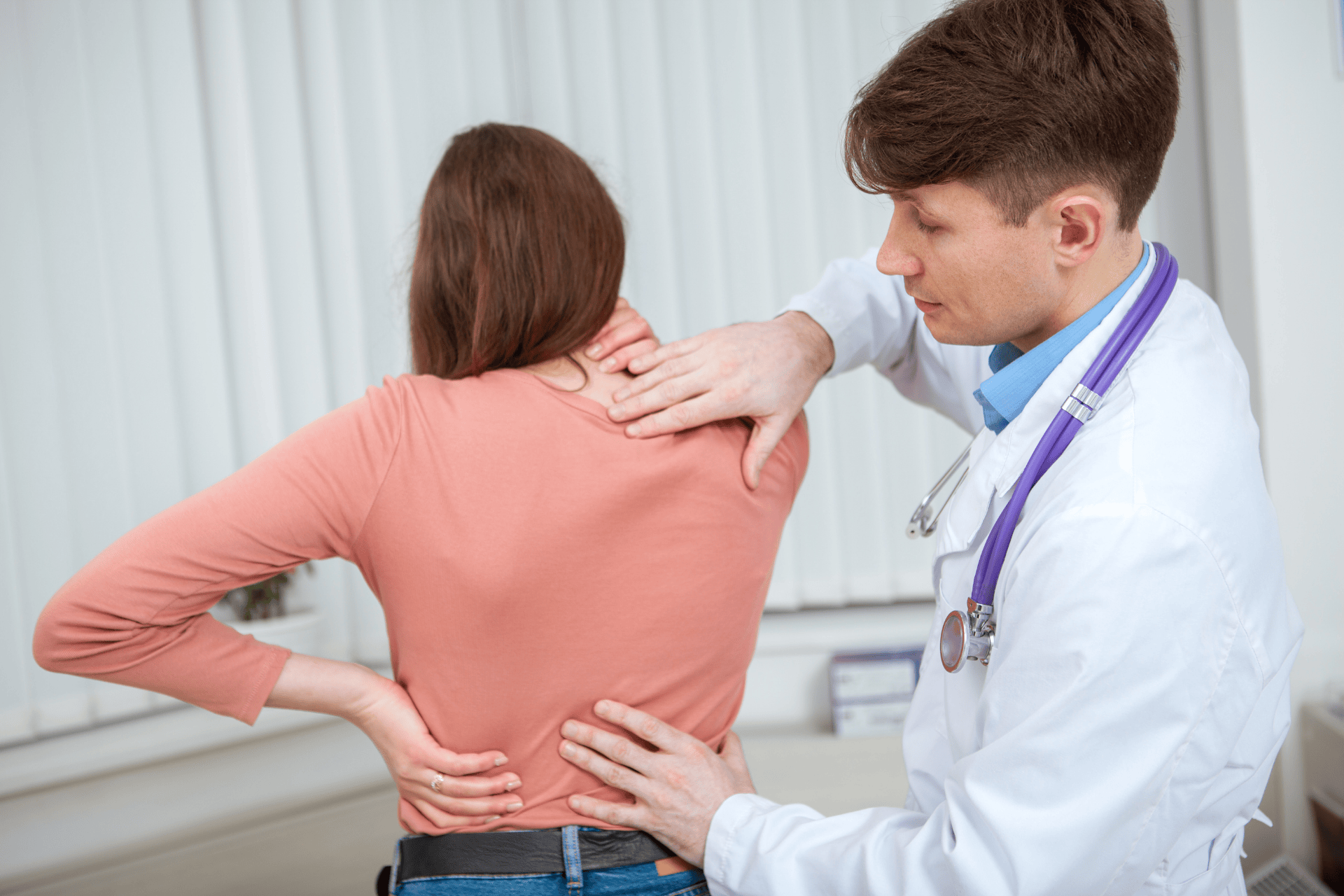 A woman with cervical pain holds her back while a doctor examines her neck and upper spine in a clinic.