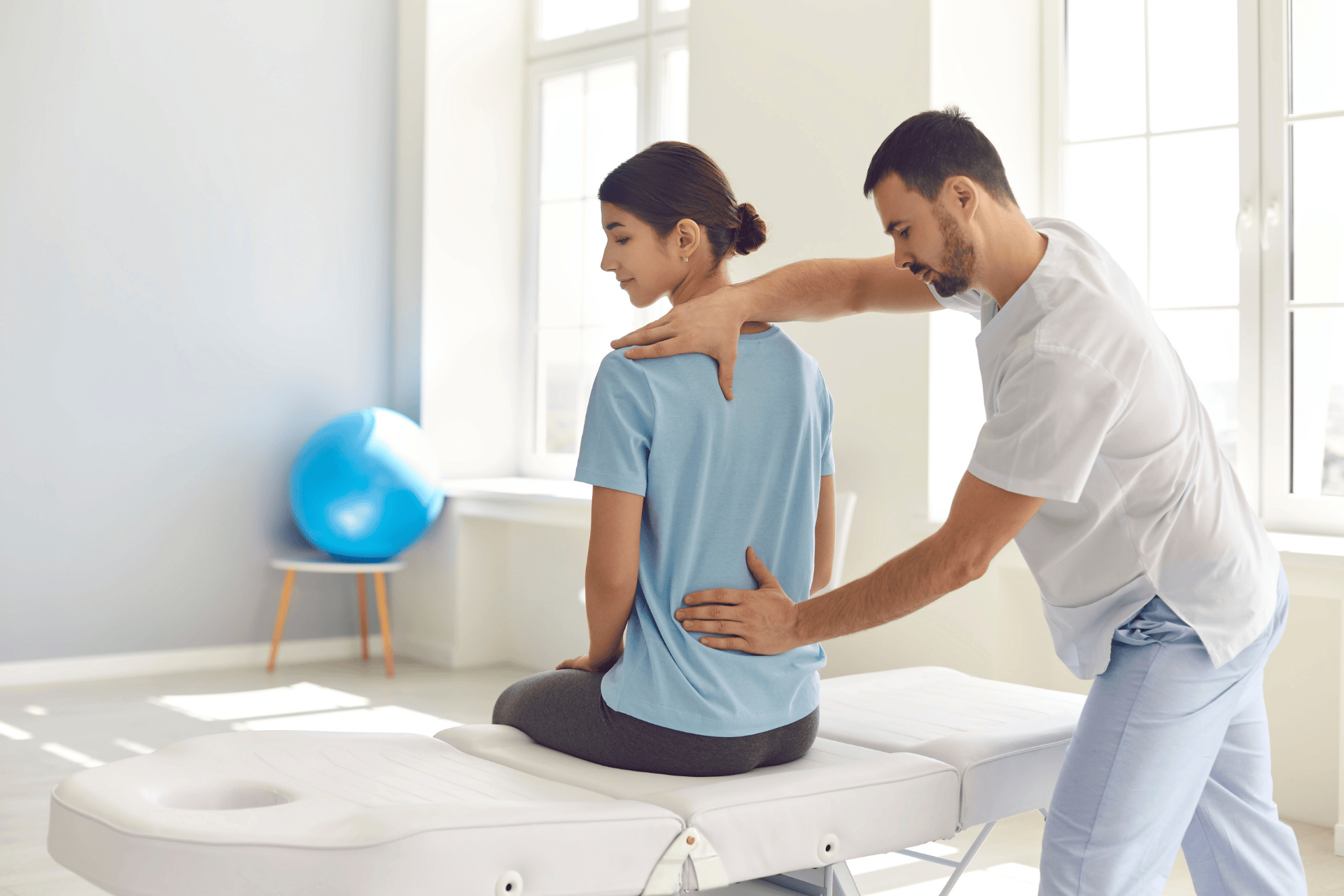 A woman sits on an exam table while a healthcare provider places hands on her upper and mid-back, indicating thoracic spine pain during a clinical assessment.