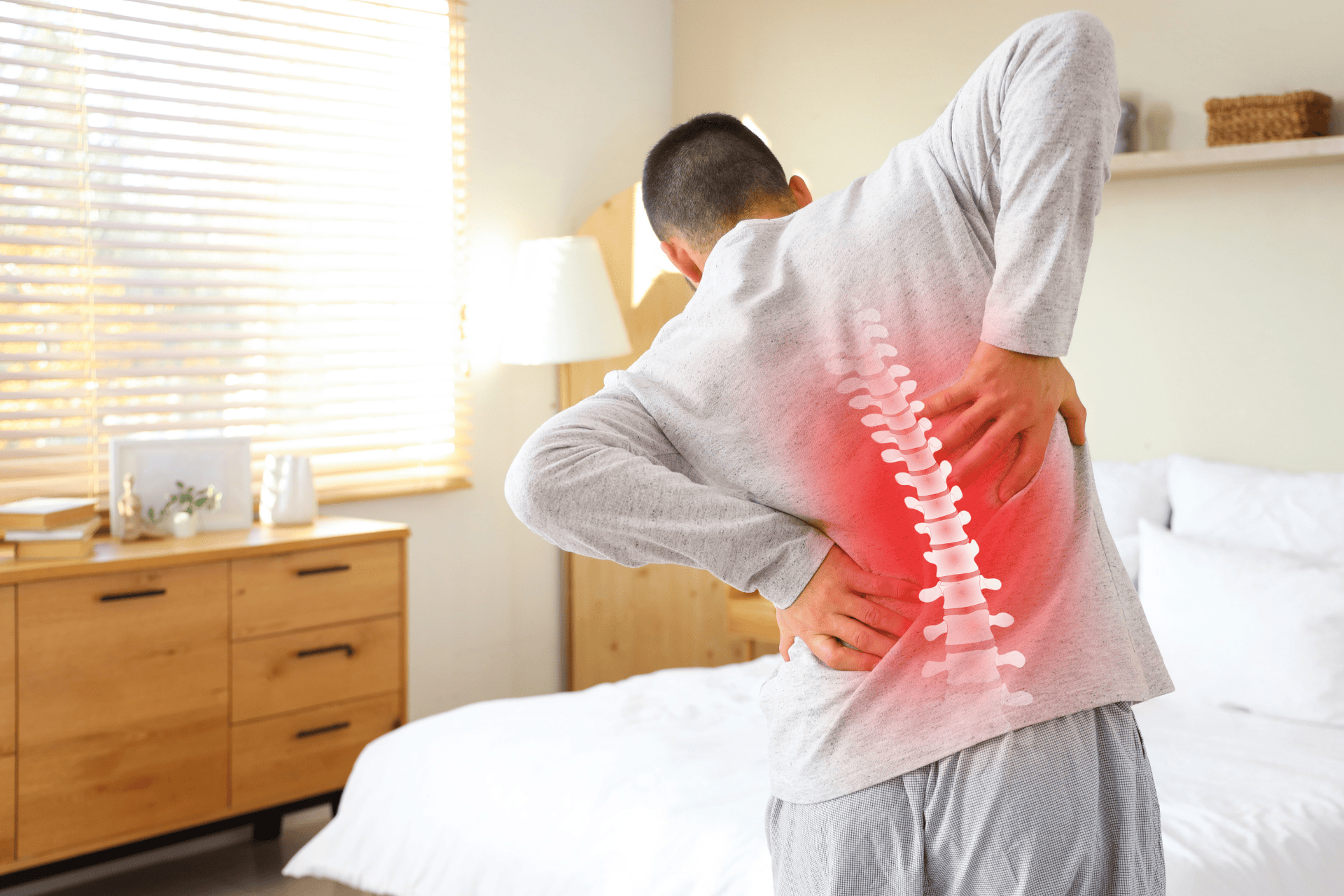 A man at home holds his lower back with both hands while a red highlighted overlay along the spine indicates back pain.