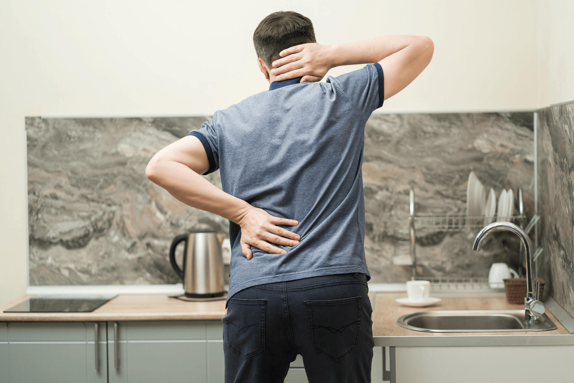 A man stands in a kitchen, holding his neck and lower back, suggesting spinal arthritis and stiffness.