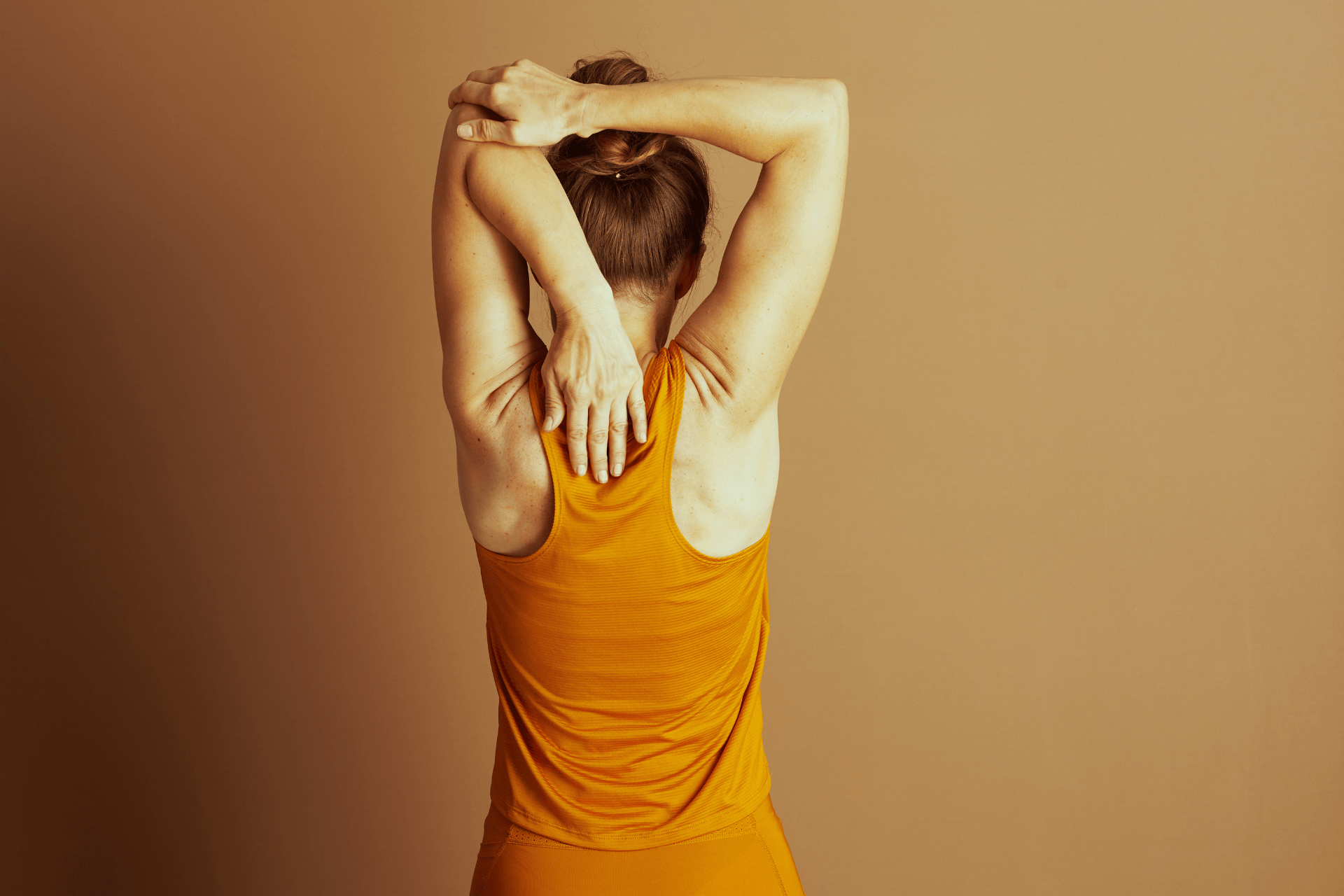 A woman seen from behind stretches her upper back and shoulders by raising one arm overhead and pressing the opposite hand between her shoulder blades.