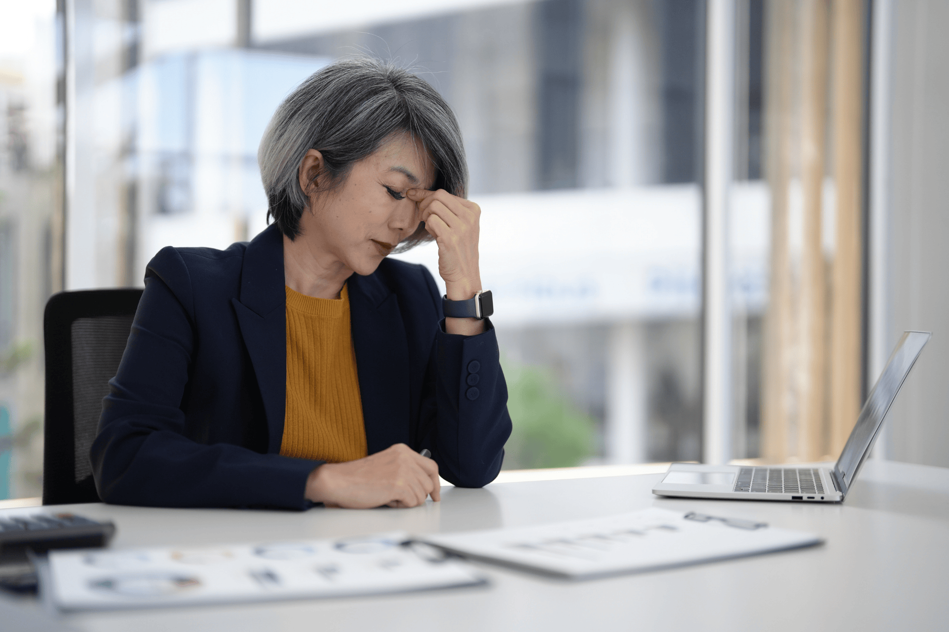 A woman sits at a desk with her eyes closed and her hand on her forehead, showing fatigue and exhaustion associated with perimenopause.