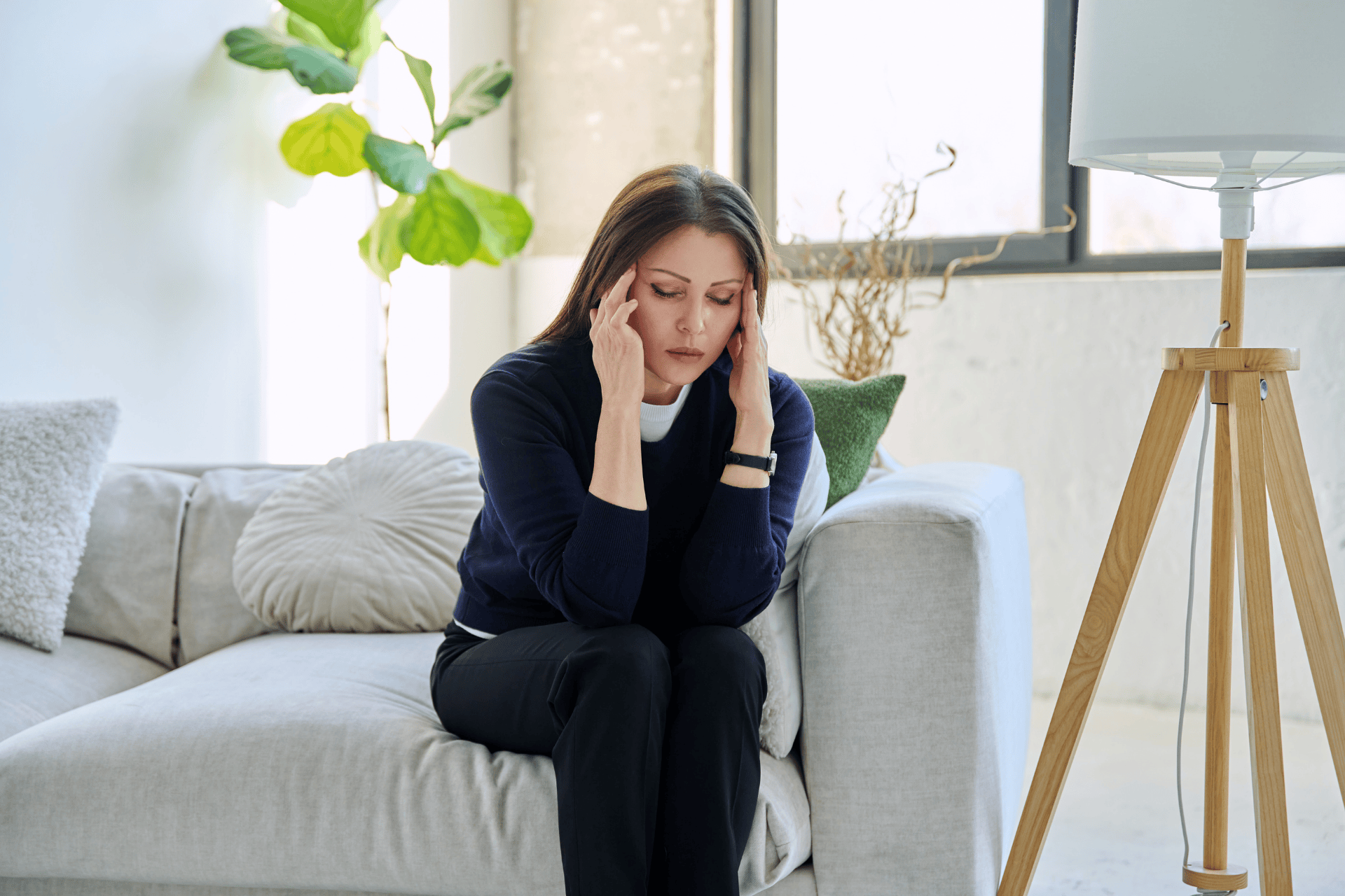 A woman sits on a light-colored sofa in a bright living room, holding her head with both hands as if stressed or experiencing a headache.