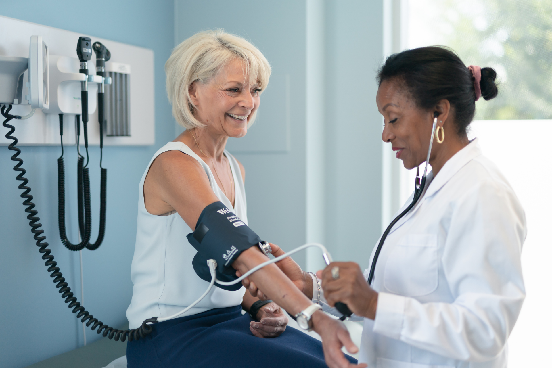 Female doctor checking a woman’s blood pressure with a cuff in a bright medical exam room, both smiling during the appointment.