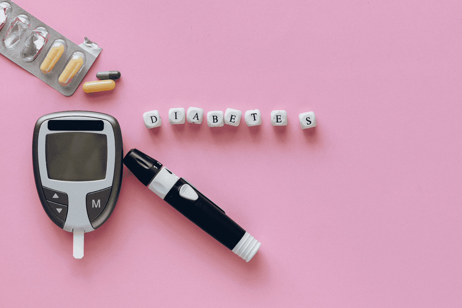 Diabetes supplies including glucometer, lancet device, pills, and word ‘DIABETES’ on pink background.