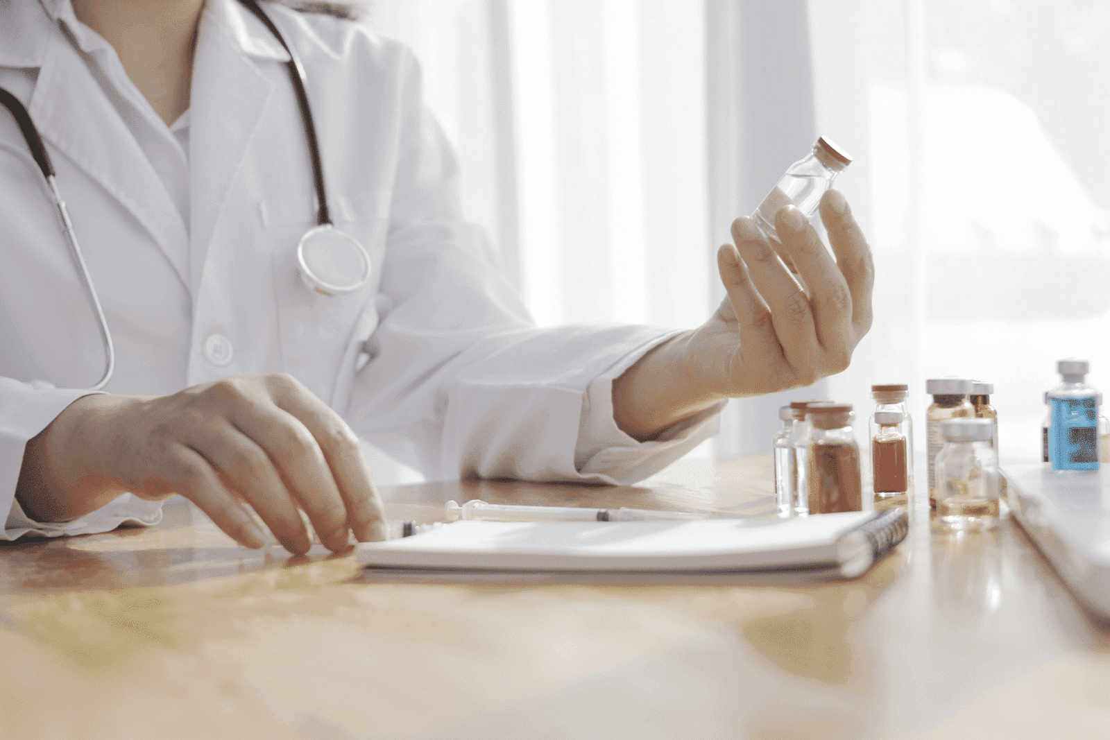 Doctor holding a small vial while sitting at a desk with several medicine bottles and a notepad.