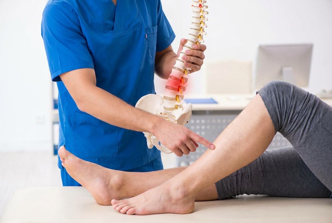 Doctor in blue scrubs holds a spine model with a red inflamed area, pointing to a patient's leg.