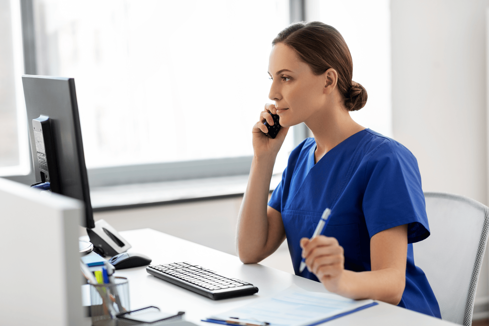 Doctor in blue scrubs talking on the phone at a desk with a computer