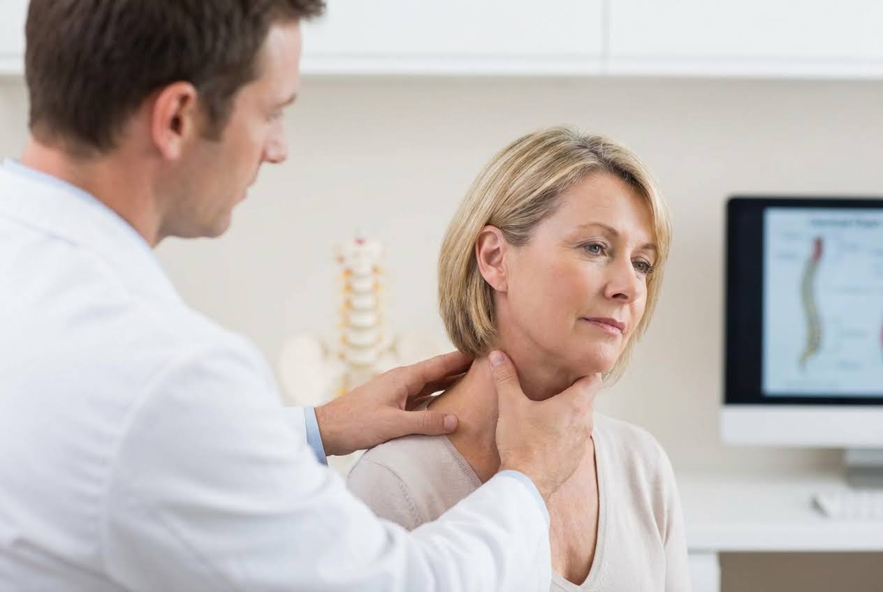 Doctor in a white coat examining a blonde woman's neck, with a spine model and computer screen in the background