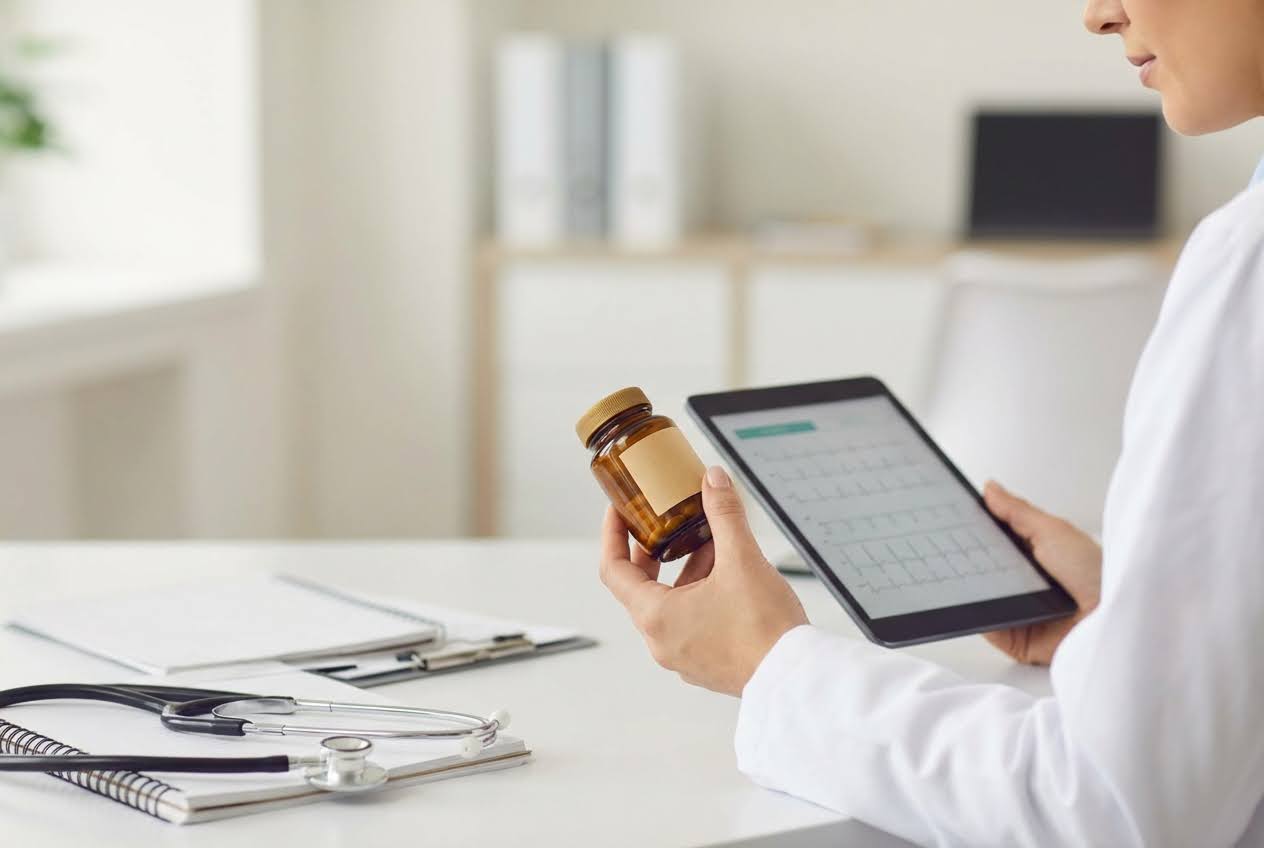 Doctor in a white coat holding a brown medicine bottle and a tablet displaying medical data, with a stethoscope on the desk