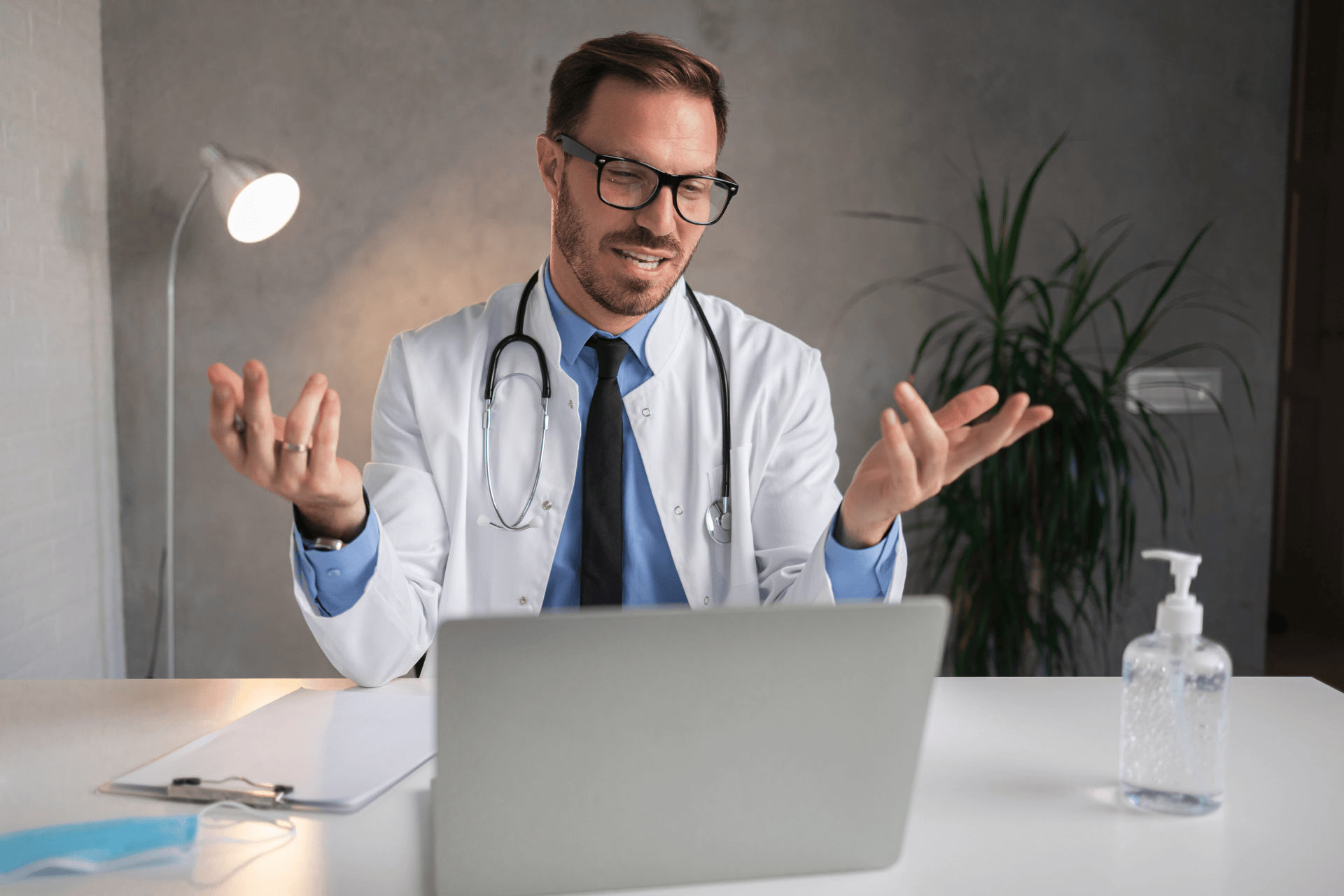 Doctor with glasses gesturing while talking during an online consultation on a laptop.