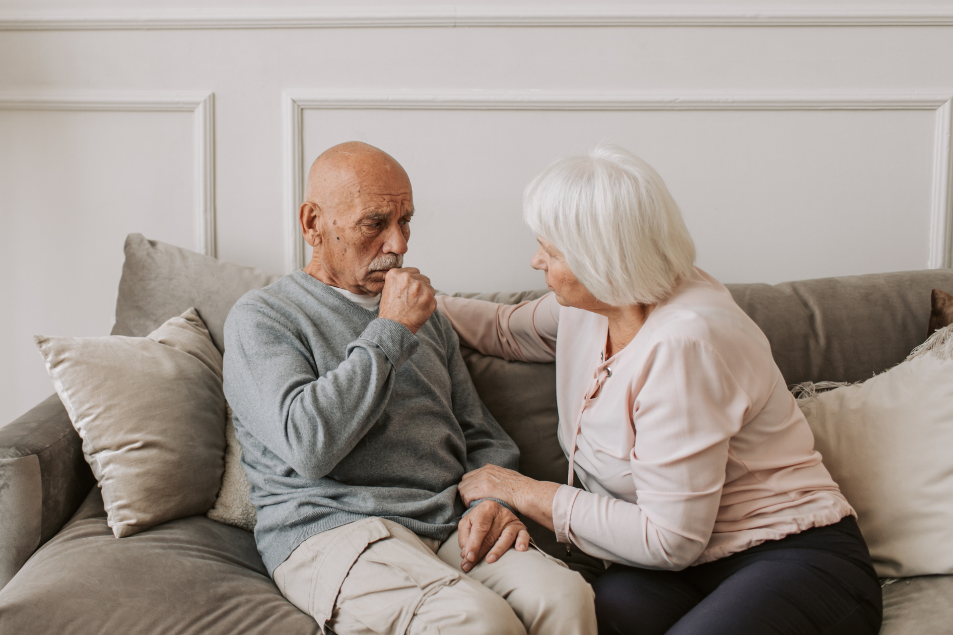 An elderly man sitting on a couch, coughing while an older woman sits beside him, offering comfort and support
