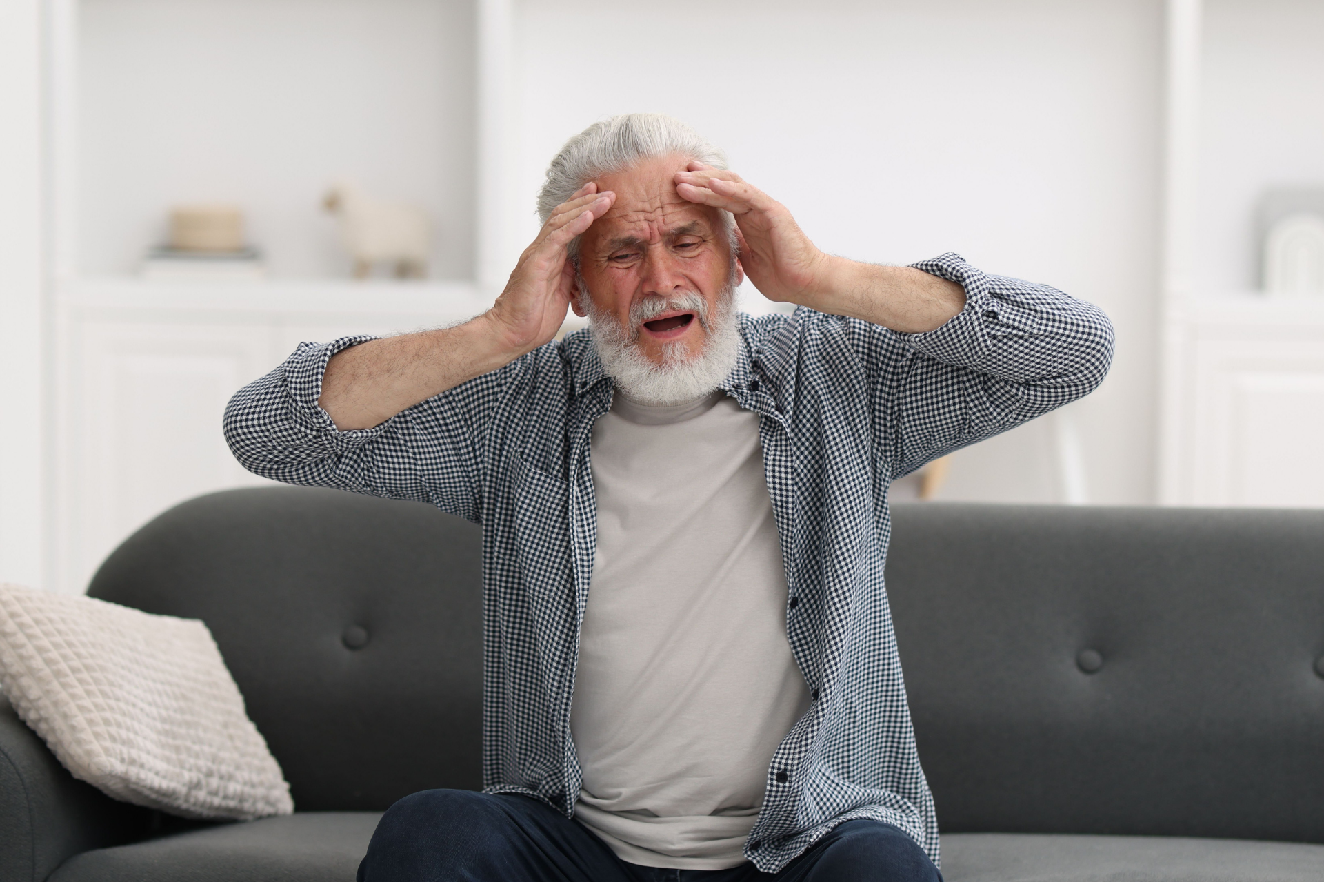 Elderly man sitting on a couch holding his head, appearing dizzy or in pain at home