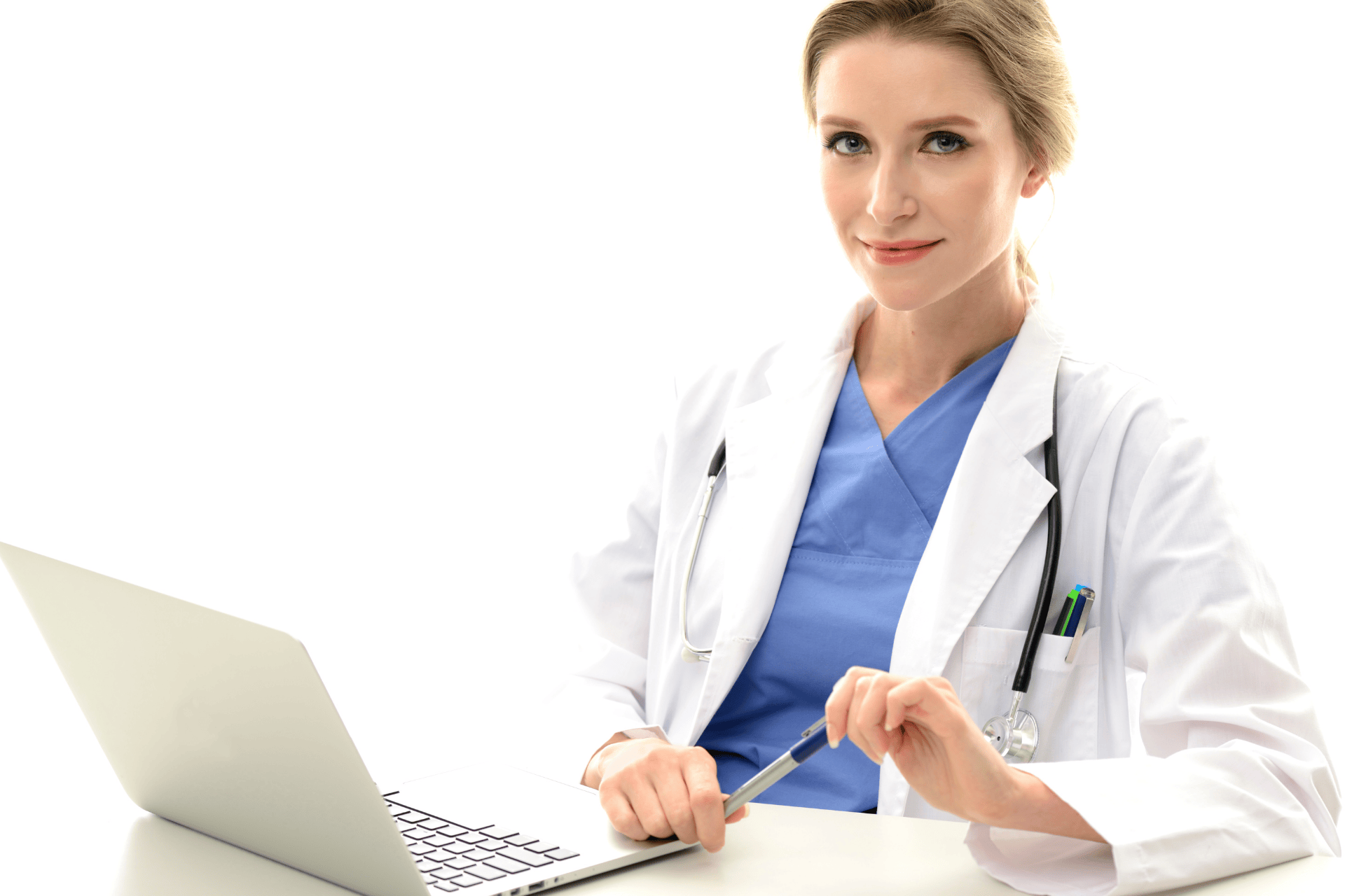 Female doctor in a white coat and blue scrubs sitting at a desk with a laptop, smiling confidently while holding a pen.