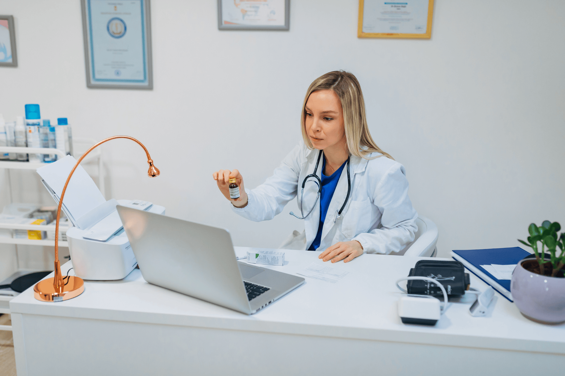 Female doctor in a white coat holding a small vial while looking at a laptop.