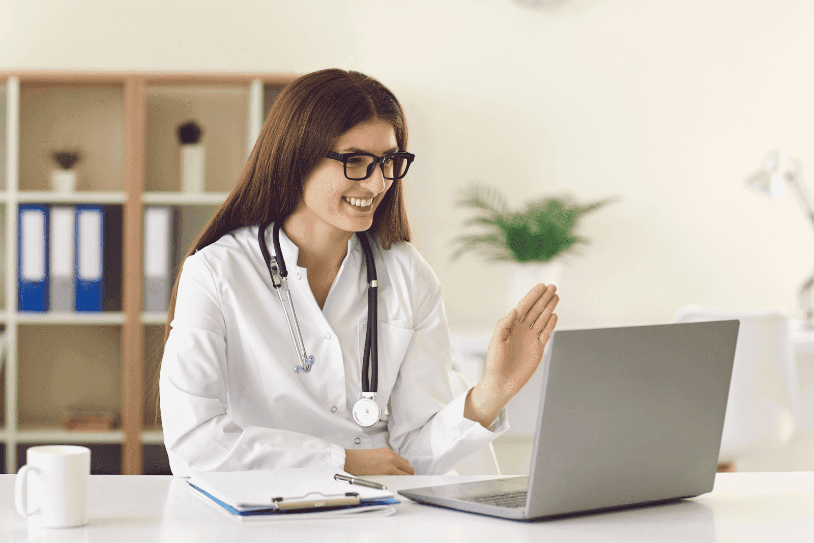 Female doctor smiling and waving during a video call on a laptop.