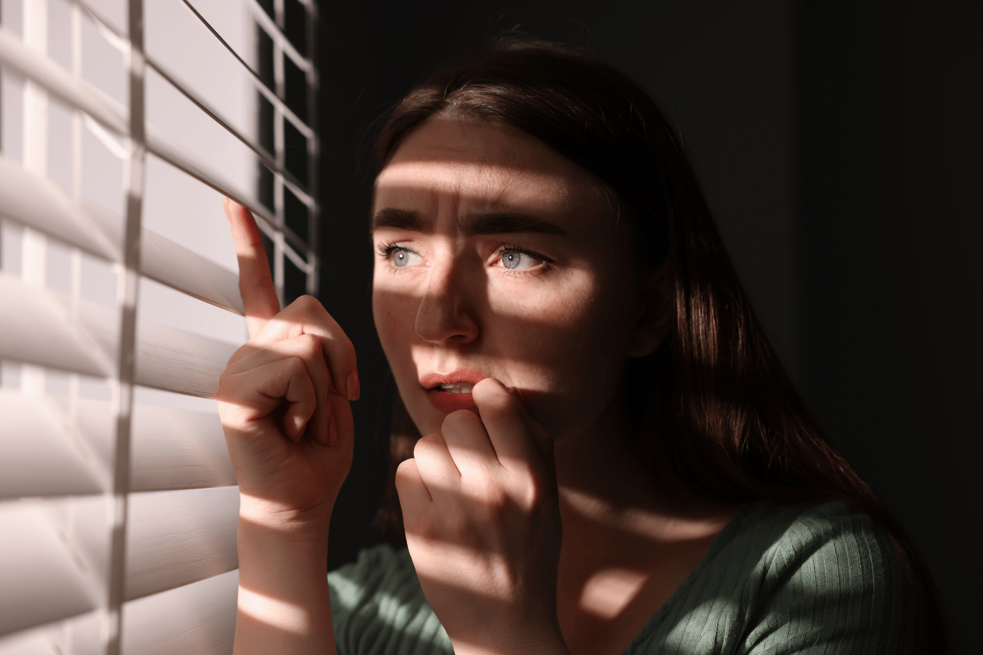 Worried woman peeking through window blinds with sunlight casting shadows on her face.