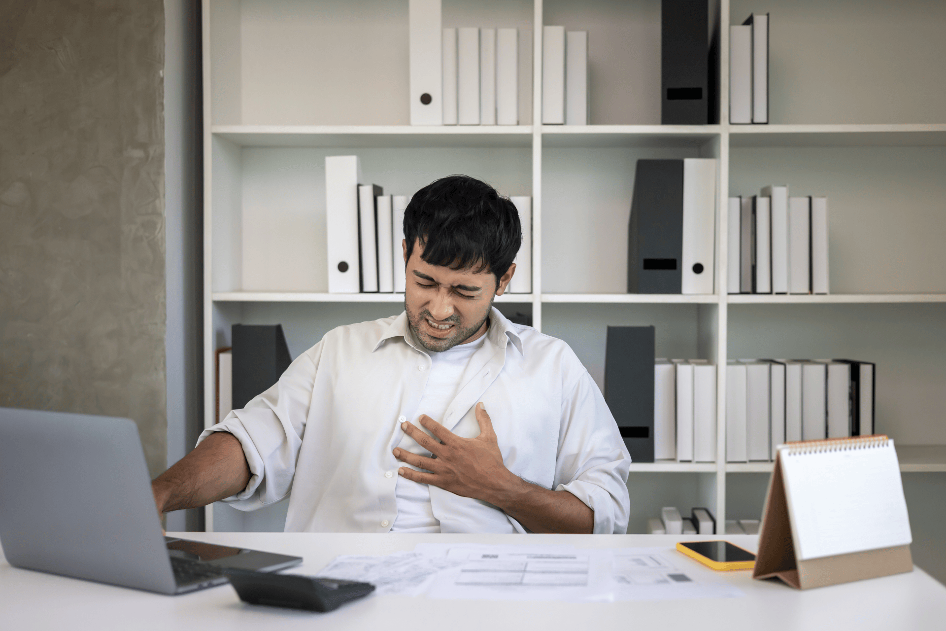 Man sitting at a desk, clutching his chest in pain while working on a laptop.