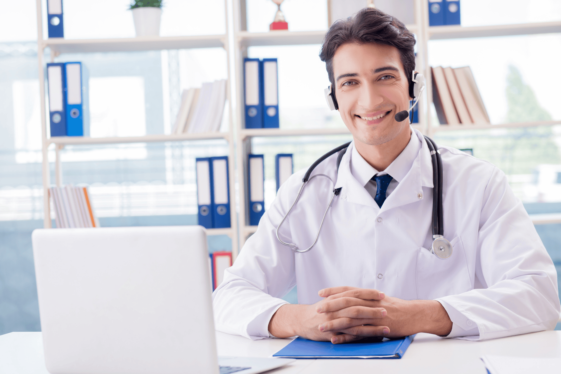 Smiling doctor wearing a headset and stethoscope, sitting at a desk with a laptop.