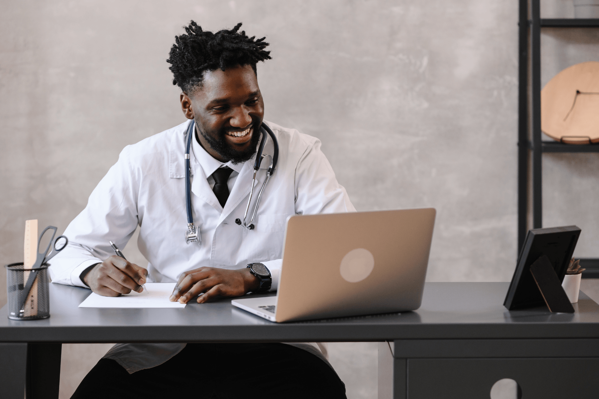 Smiling doctor sitting at a desk, writing notes while looking at a laptop.