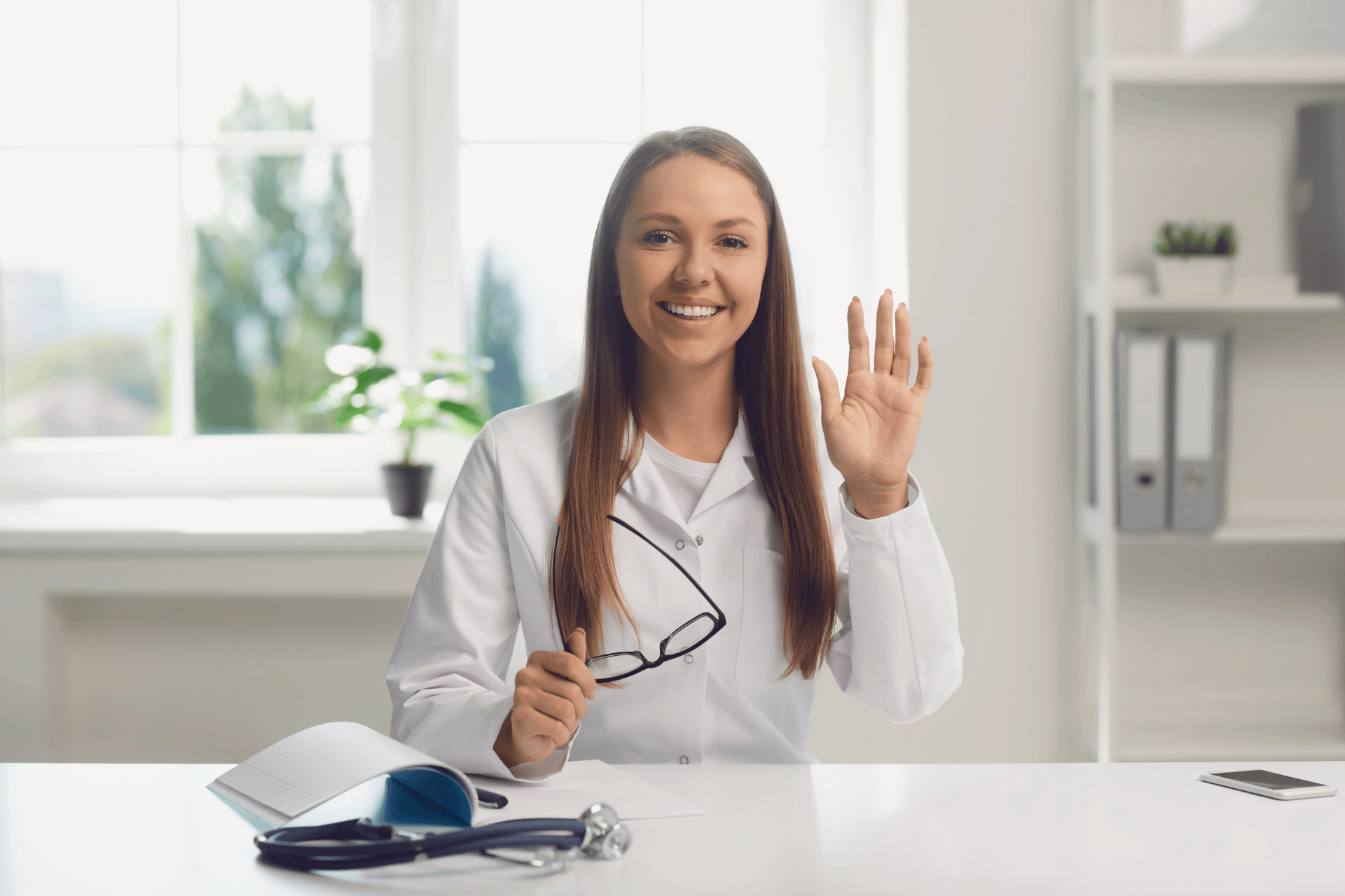 Smiling doctor waving while sitting at a desk with medical tools.