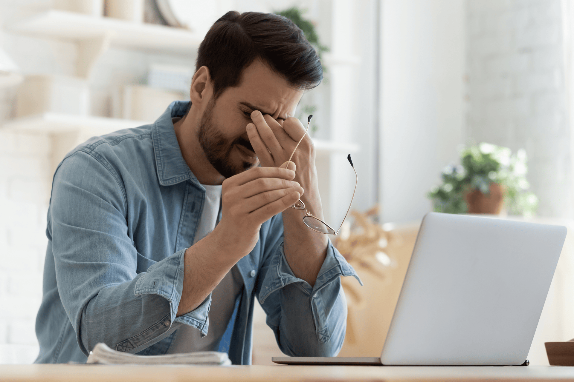 Man sitting at desk rubbing his eyes in frustration while working on a laptop.